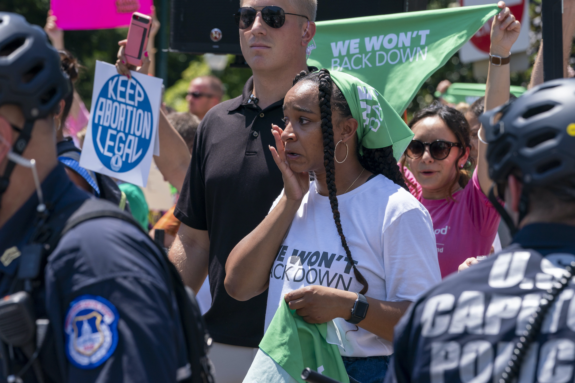 A person tearing up in a crowd of people. The person, who is holding a green bandana, wears a shirt that says "We Won't Back Down." Other people hold up signs in the crowd, including "Keep Abortion Legal."