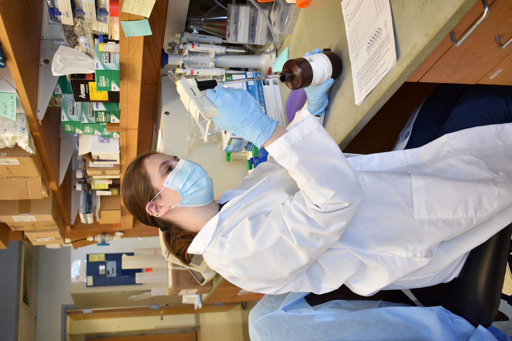 Woman with brown hair in a white lab coat pipets a clear liquid in a lab.