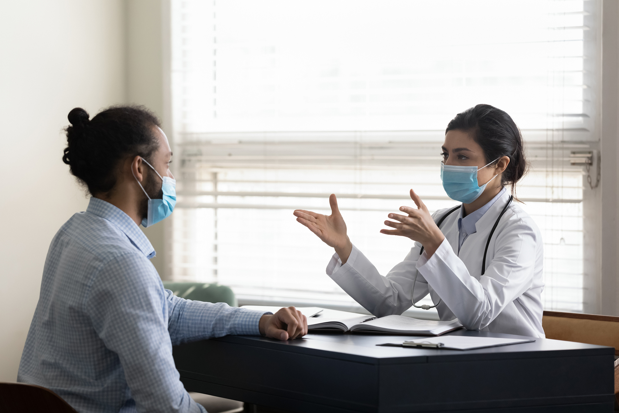 A masked doctor and patient speaking at a desk.