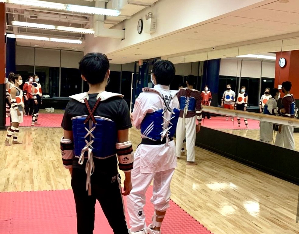 At a taekwondo practice, students in a mirrored studio stand on red mats with padding protecting their shins, forearms, and torsos.