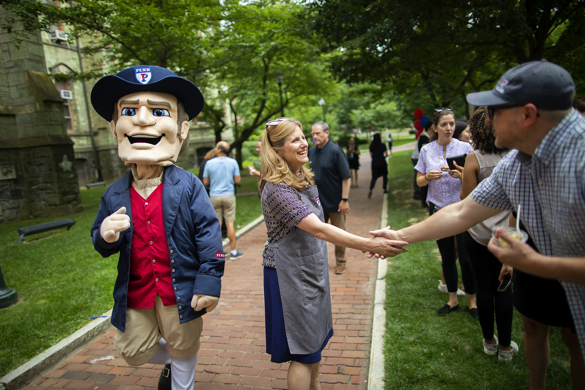 Penn President shakes hands with someone on College Green beside the Penn Quaker mascot.