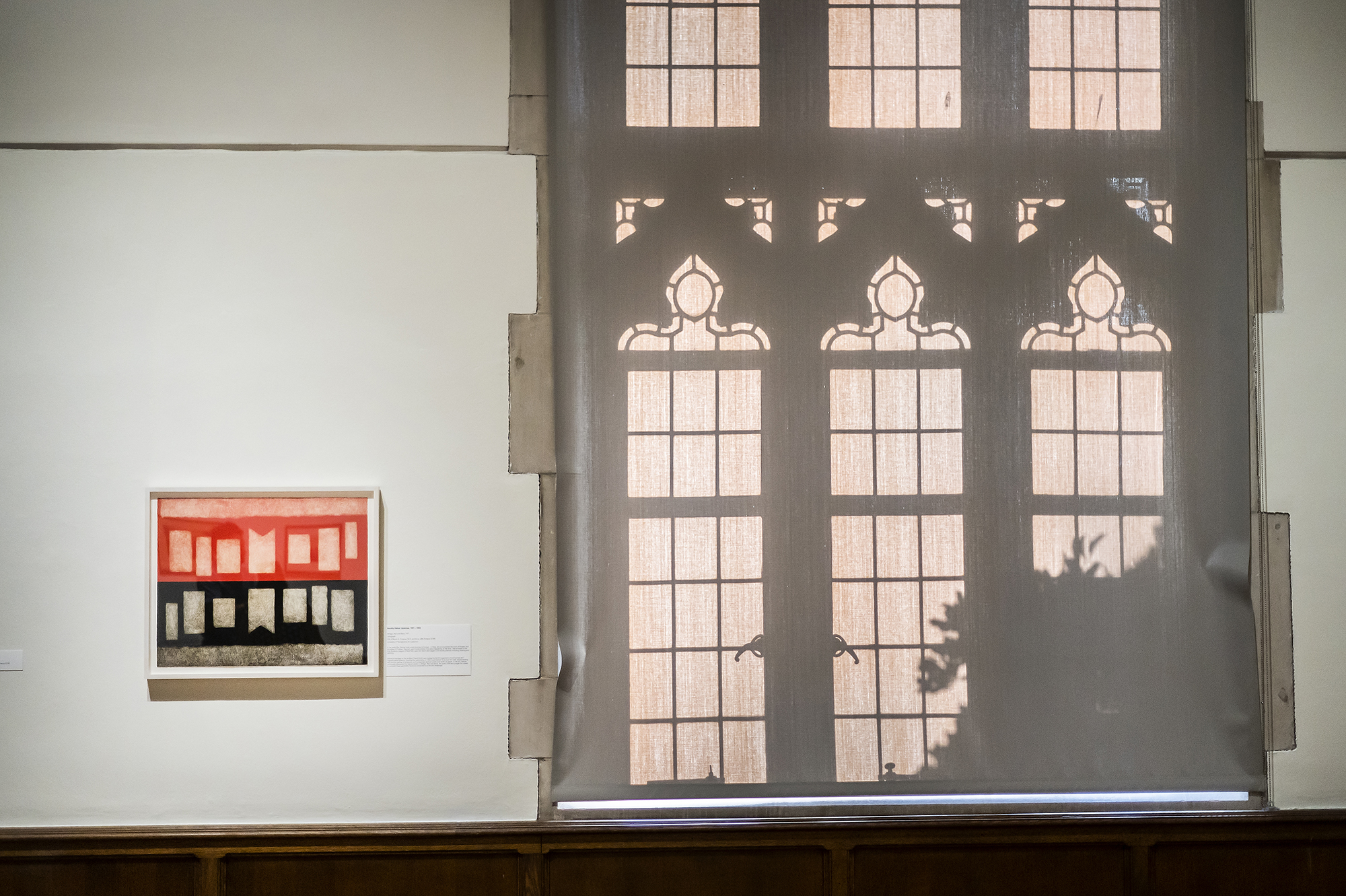 An artwork hangs on the wall beside a curtain pulled down with the reflection of ornate windows in relief.