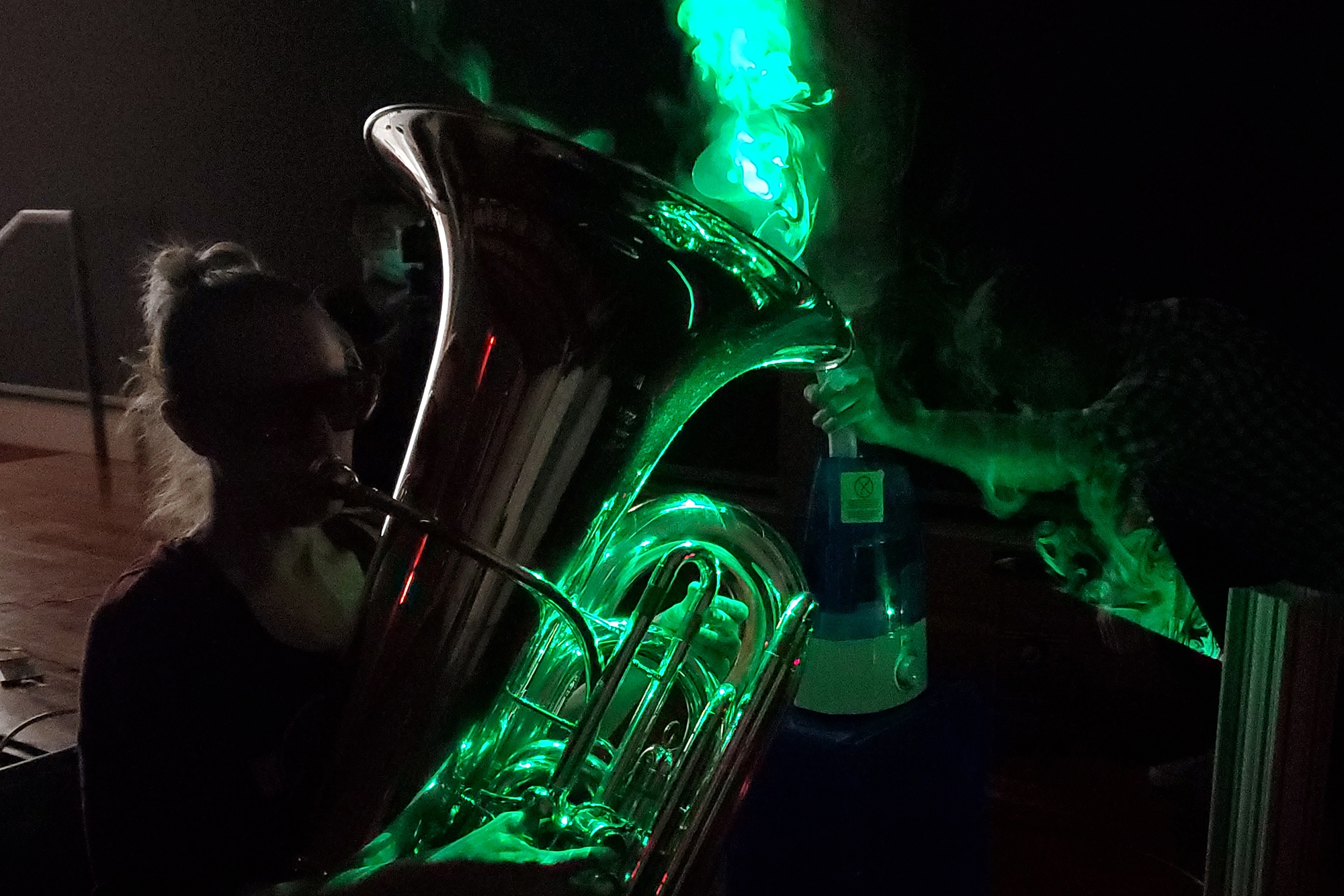 Person playing a tuba in a dark room with a green laser shedding light on water mist