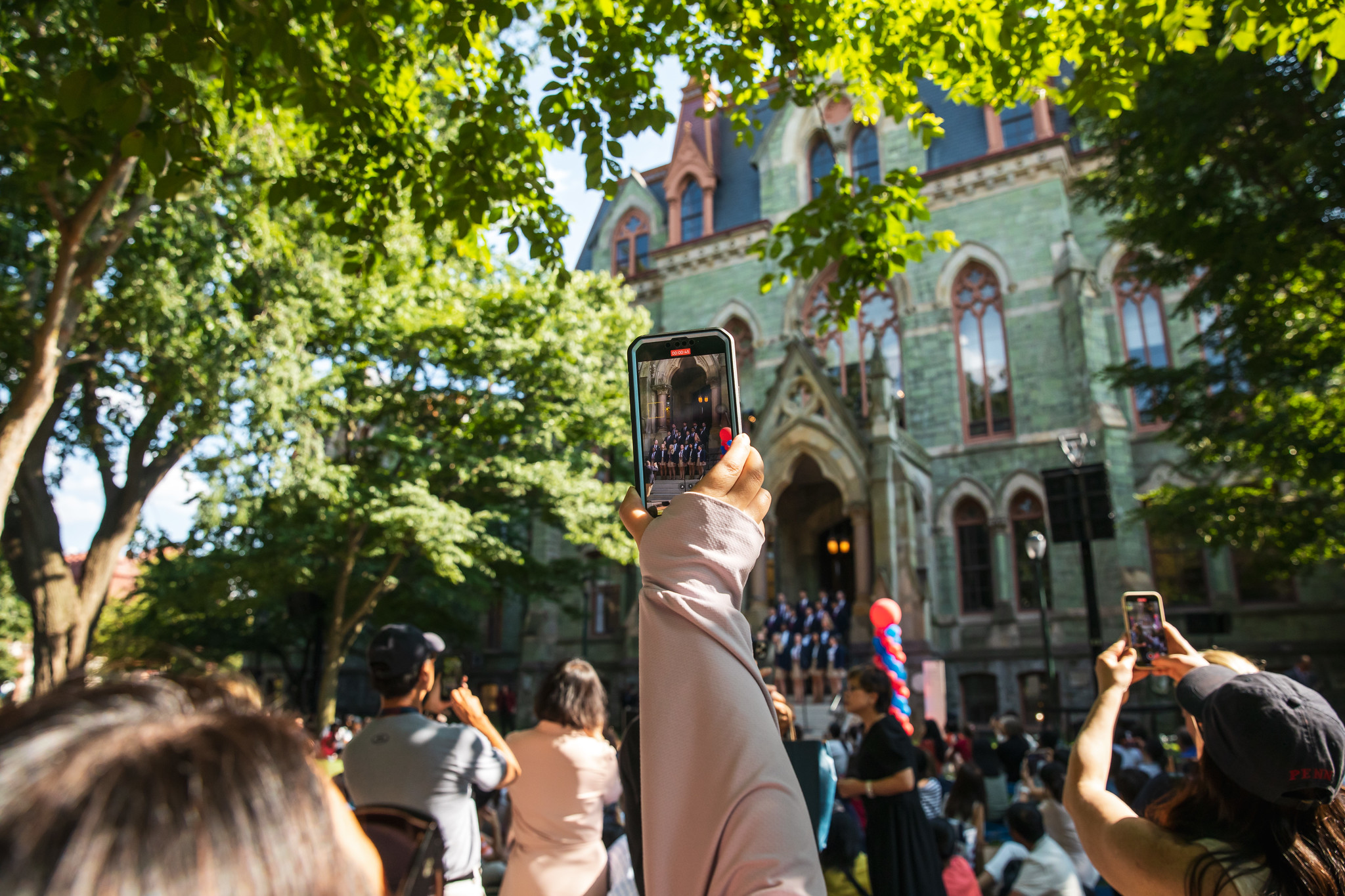 A crowd of people in front of College Hall, one holds up a smartphone to photograph a singing group on the steps.