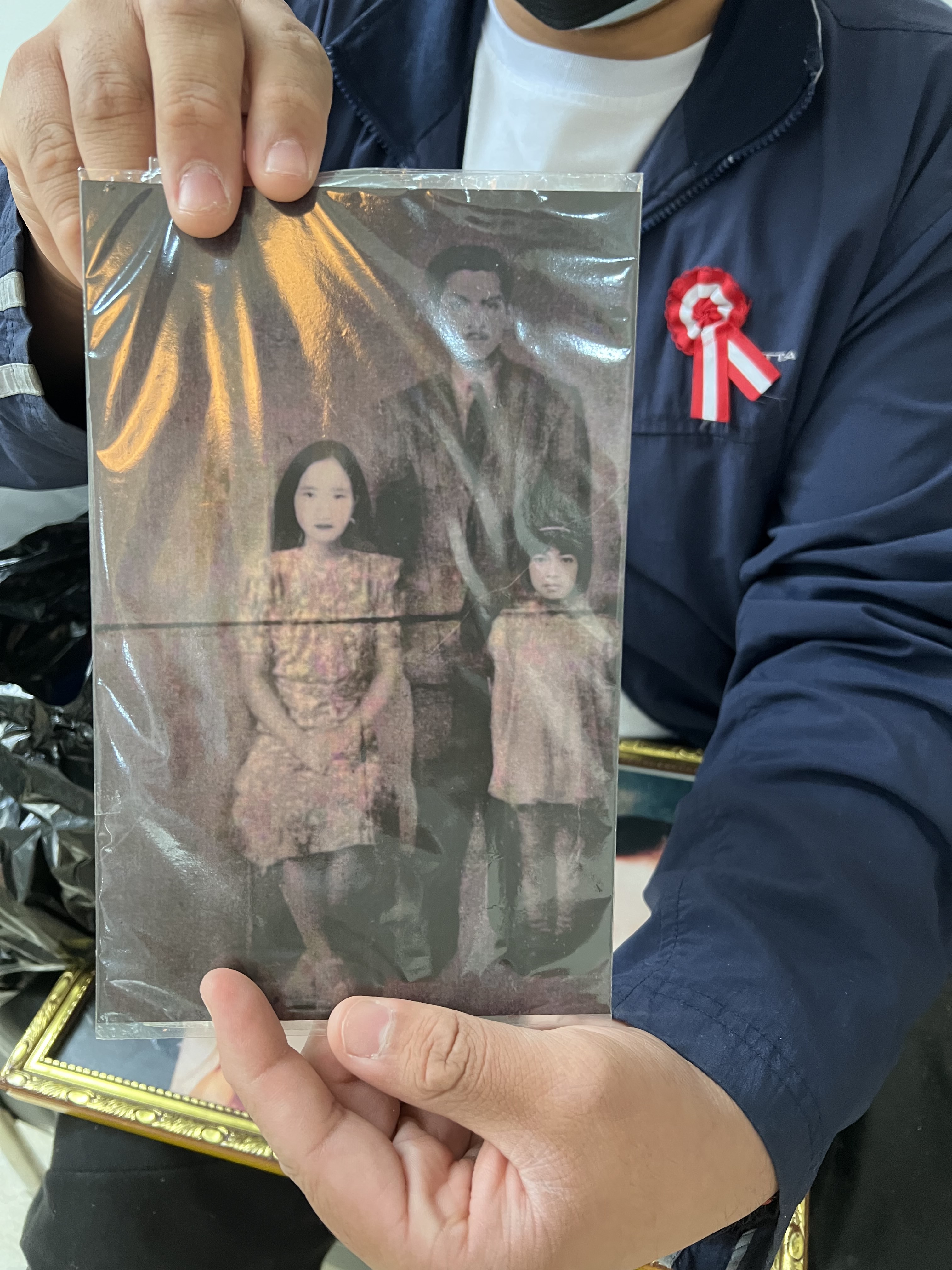 A man in a blue shirt holds up an old photograph of a man, woman, and child