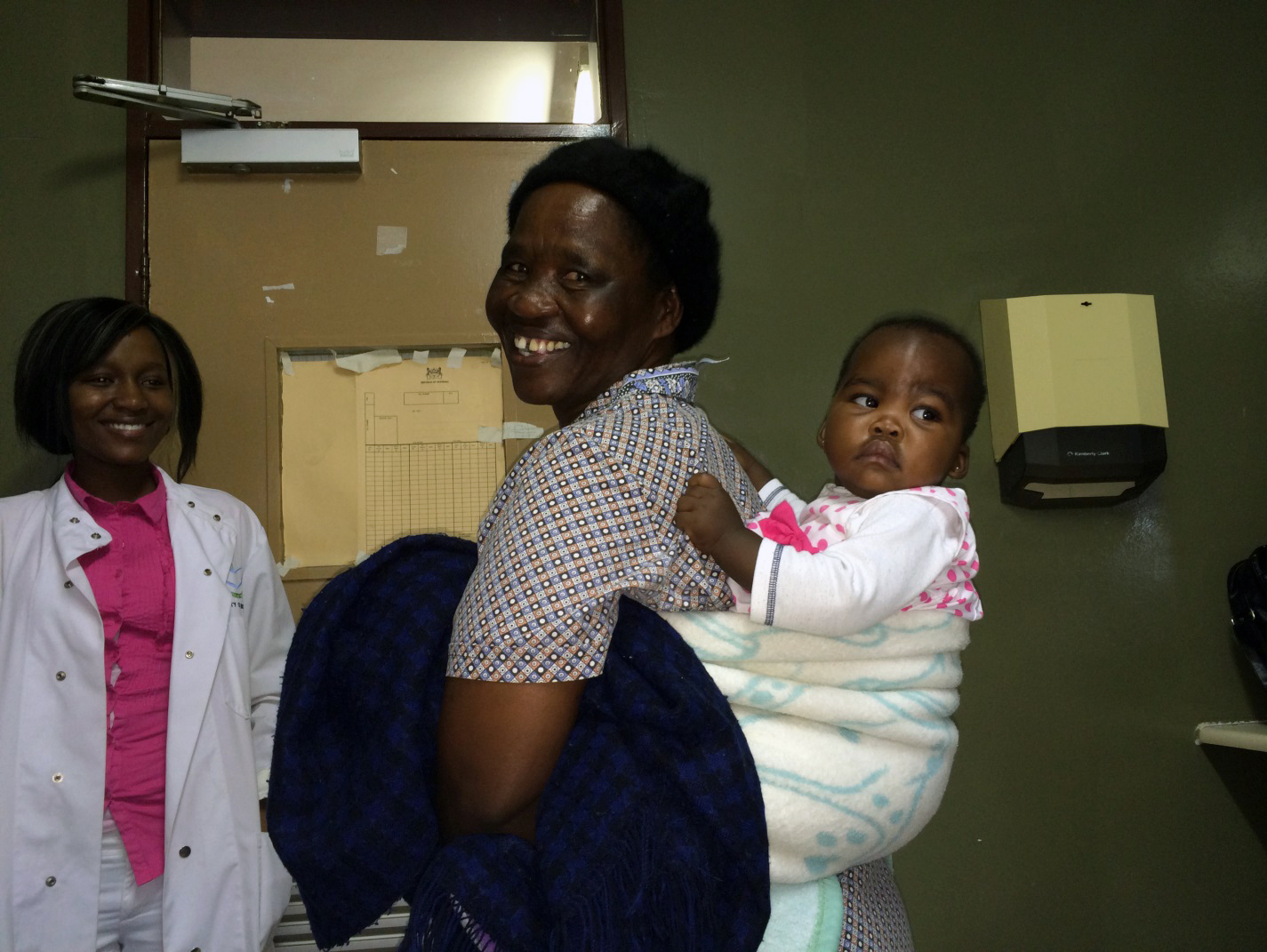 A doctor smiles at a patient carrying a baby in a baby carrier on their back.