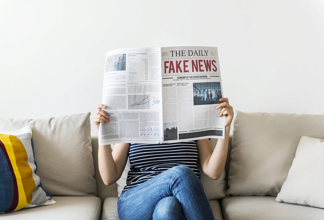 Person sitting on a couch reading a newspaper in front of their face, headline reads FAKE NEWS.