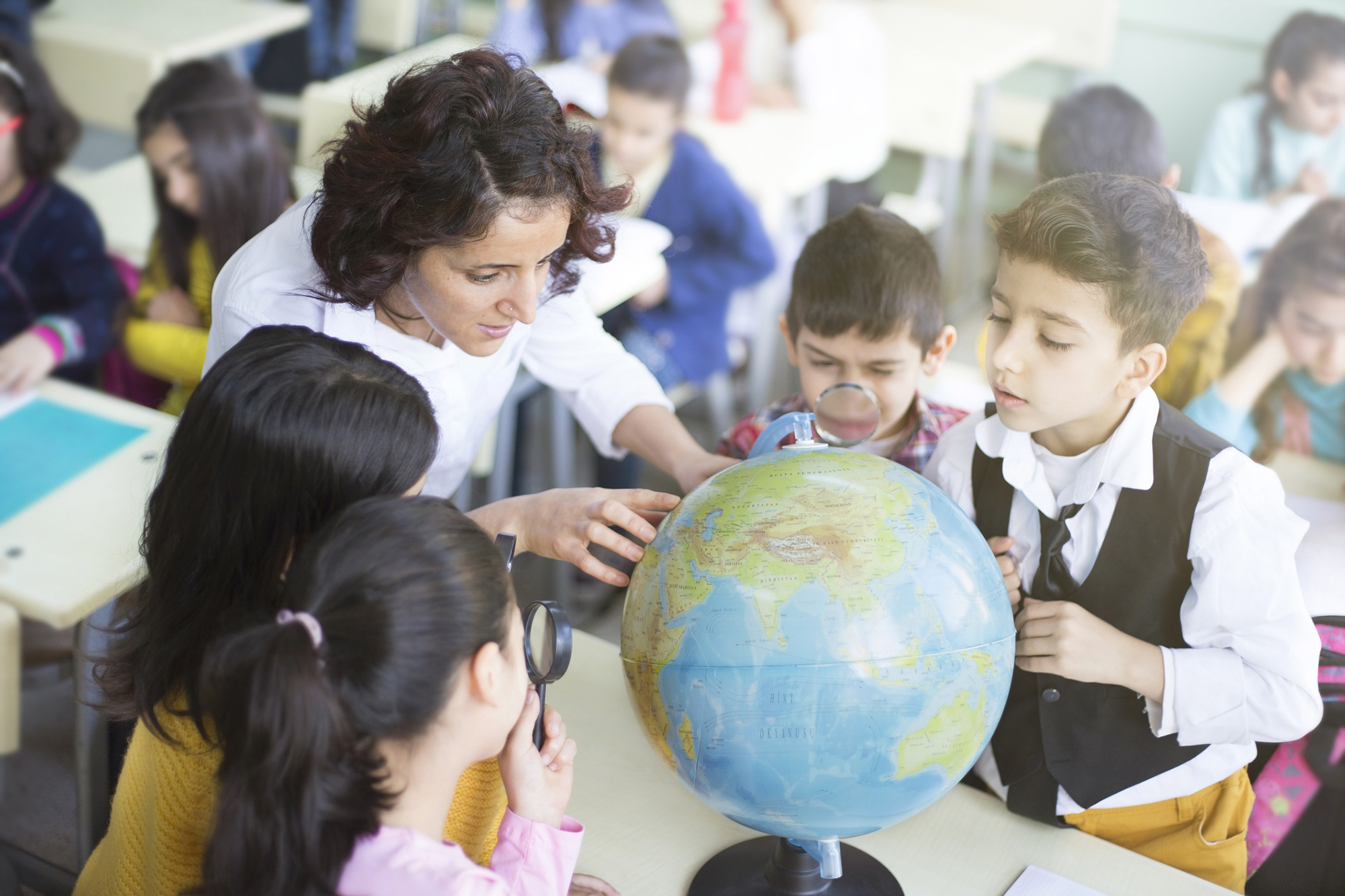 A teacher in a classroom with young children looking closely at a globe on a desk.