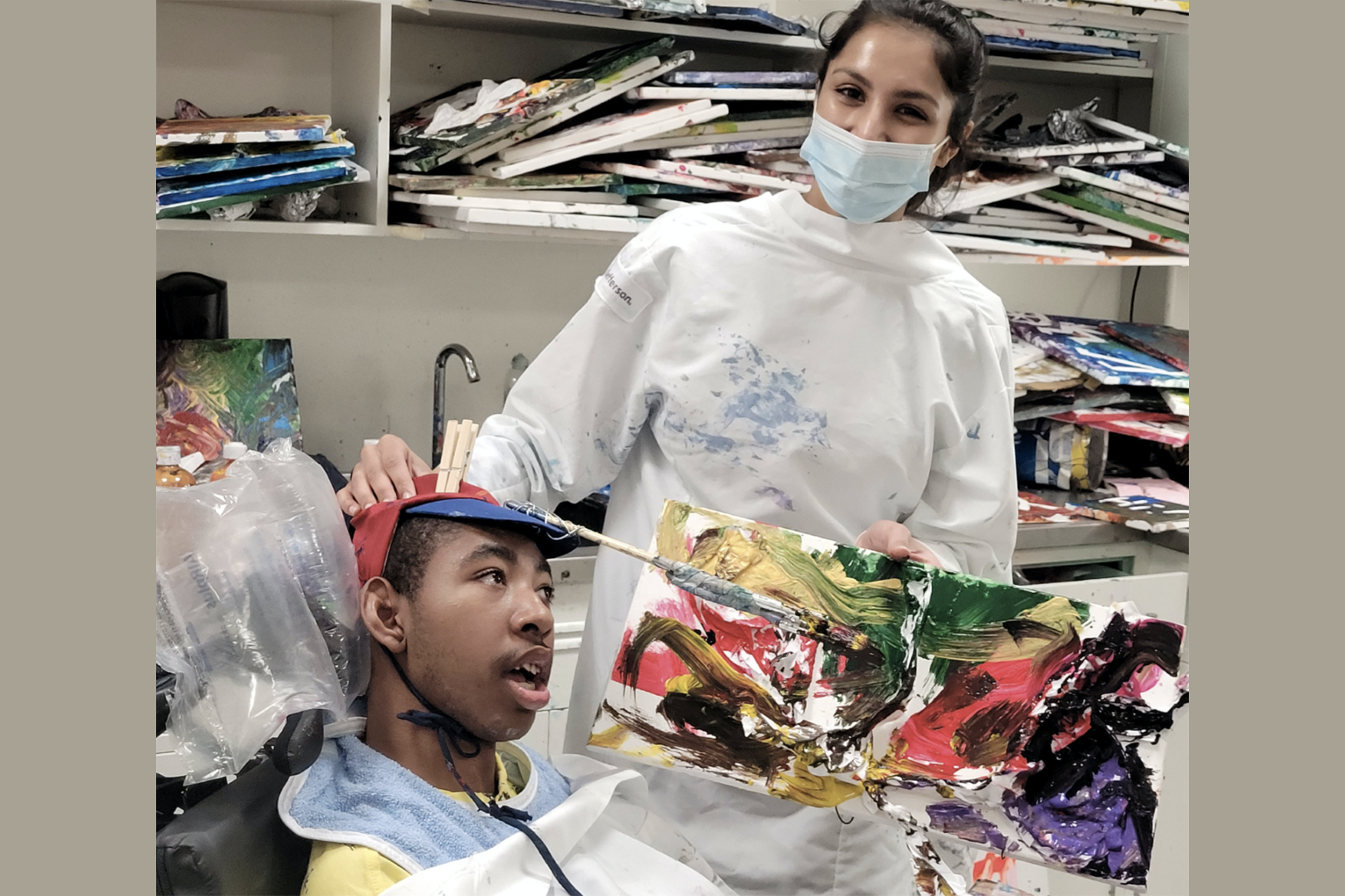 Nursing student Aman Uppal assists a student from the HMS School for Children with Cerebral Palsy during art class. She holds up a painting, and painting supplies are all around.