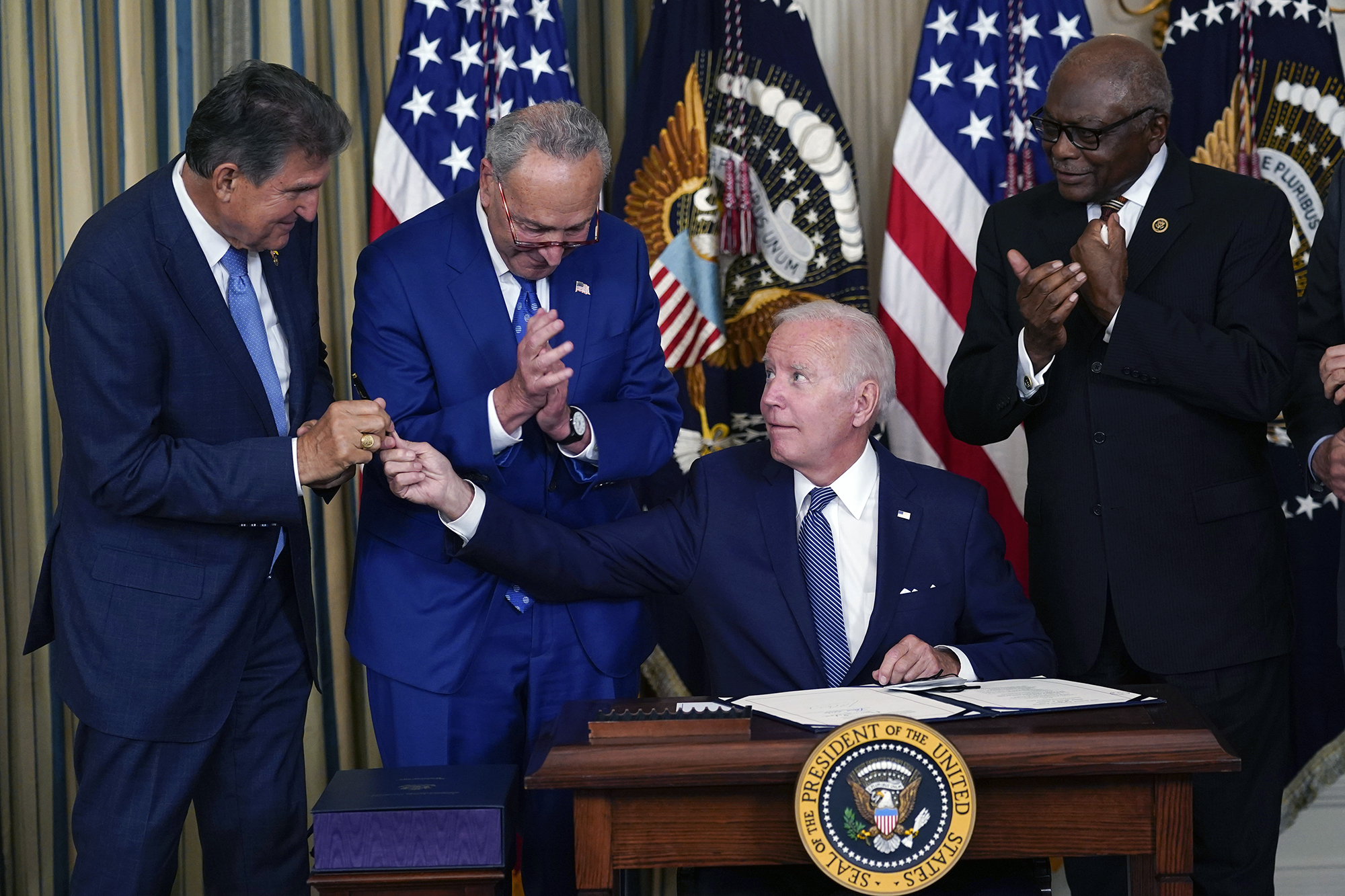 Joe Biden, Chuck Schumer, and Joe Manchin at a bill signing. Biden is sitting at a desk with the Presidential Seal. Schumer and Manchin are standing behind him. Behind all three are two American flags and a third other flag.