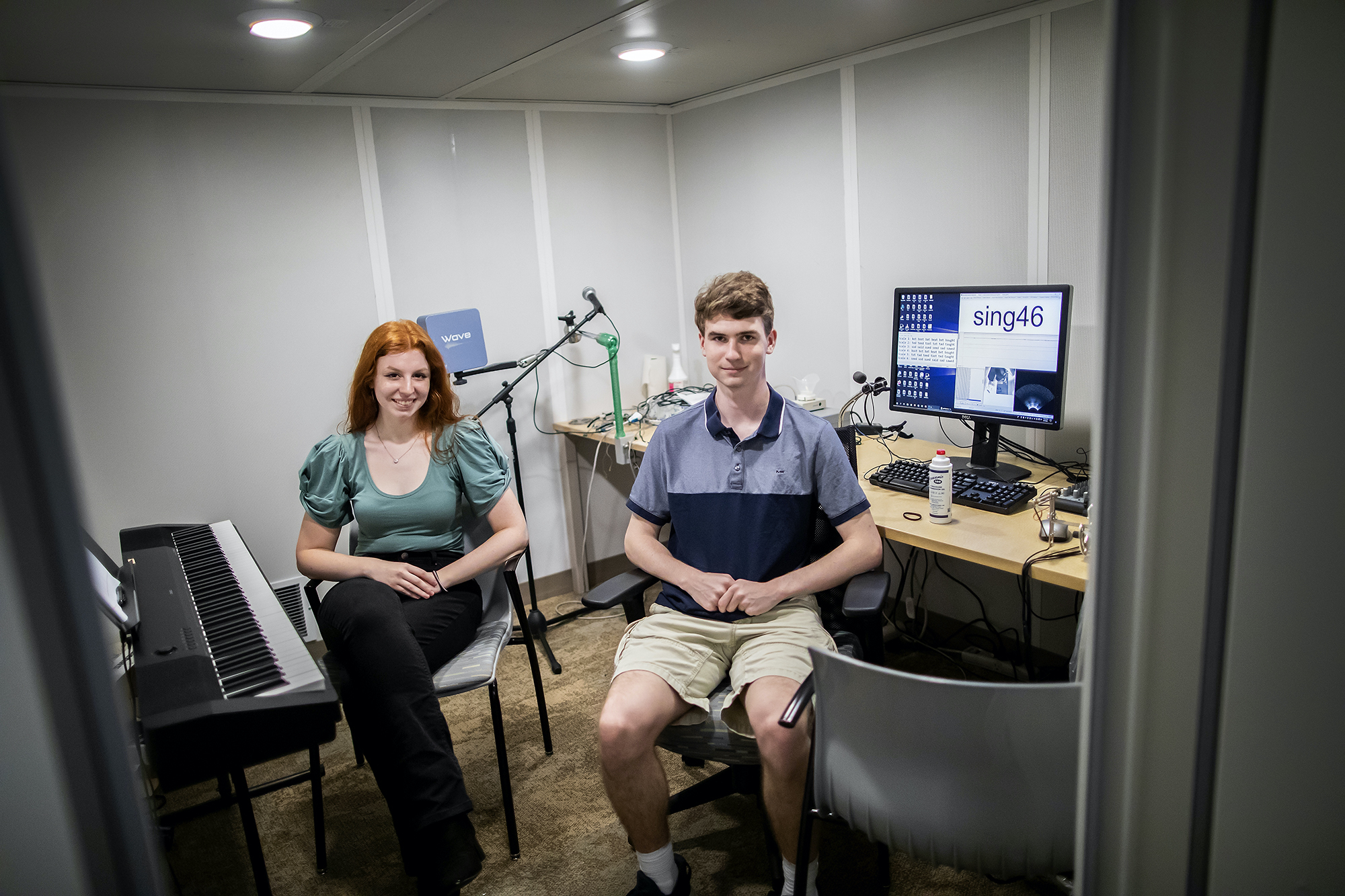 Two people sitting in a soundproof room, one next to an electric piano, a microphone behind her, the other sitting next to a computer with the words "sing46" visible on the screen, a keyboard and ultrasound gel in front.