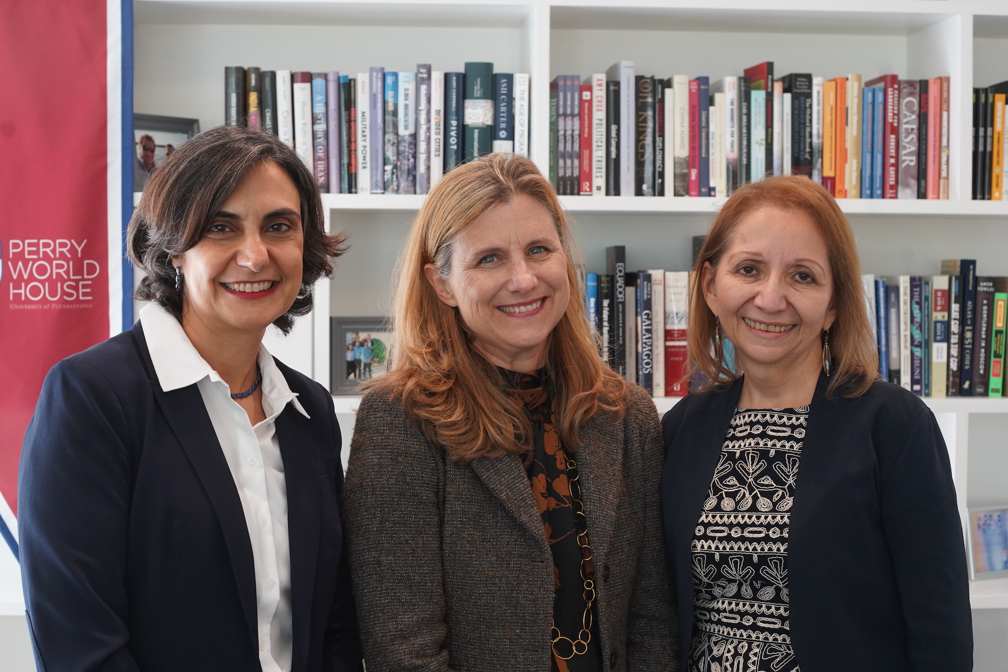 Tulia Falleti, Liz Magill, and Antonia Villarruel pose in front of bookshelves. A banner in the back reads: Perry World House. 