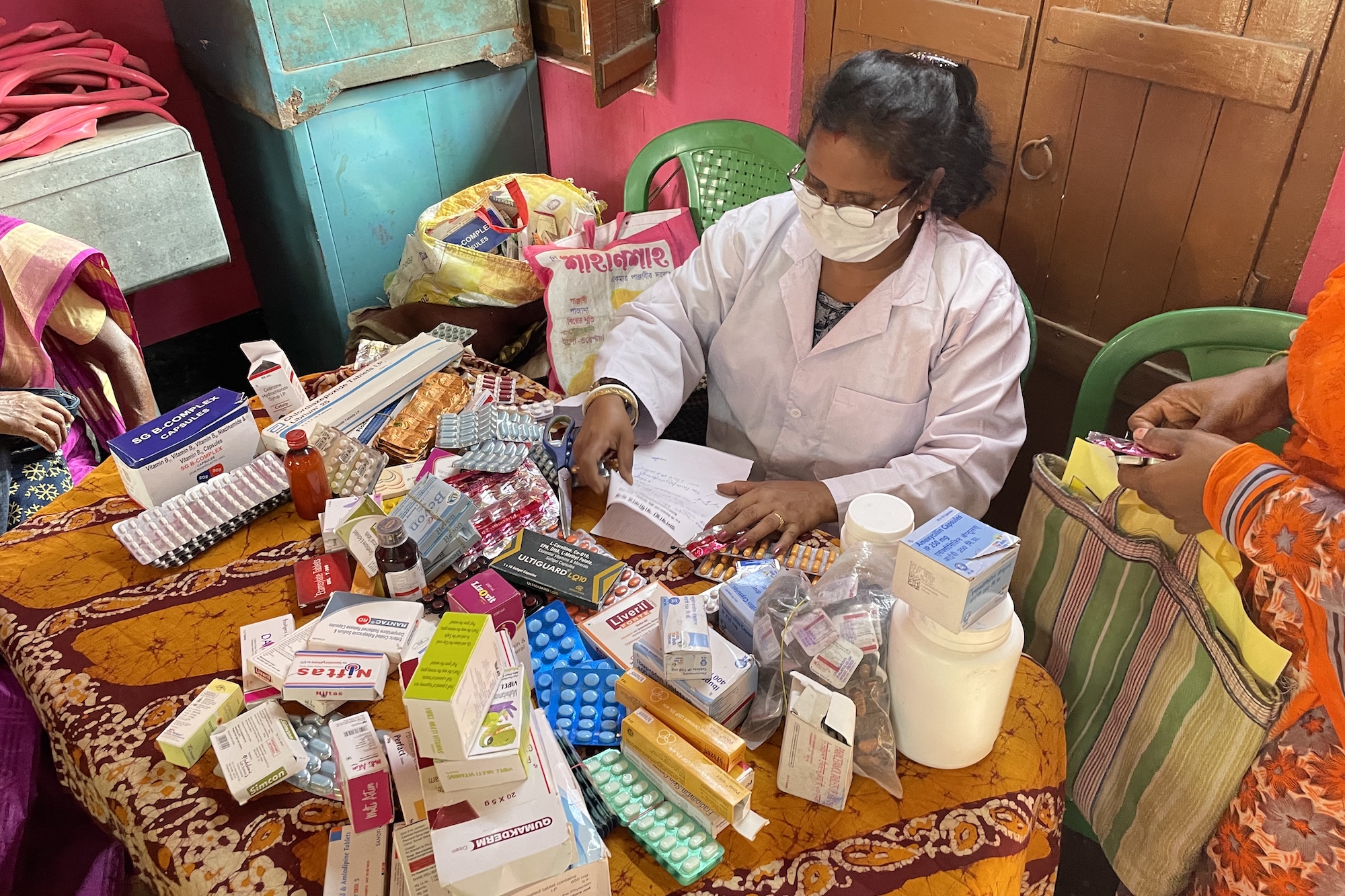 A woman wearing a mask and a white lab coat sits at a table covered in a colorful tablecloth, pills, and packages of medication