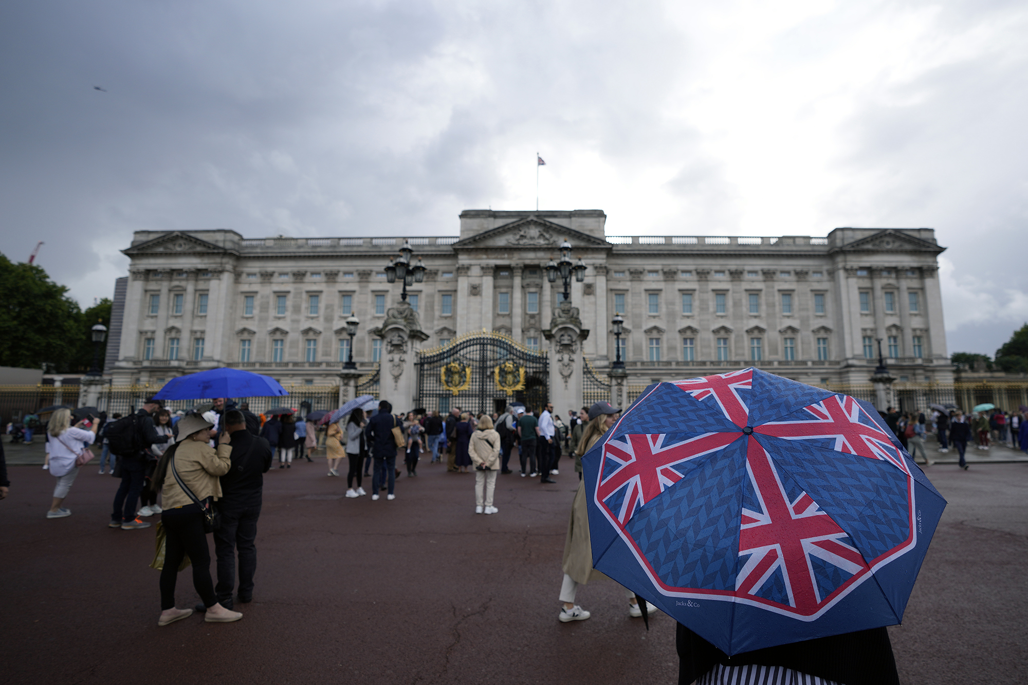 Buckingham Palace in background, people standing outside the gates, some holding umbrellas.