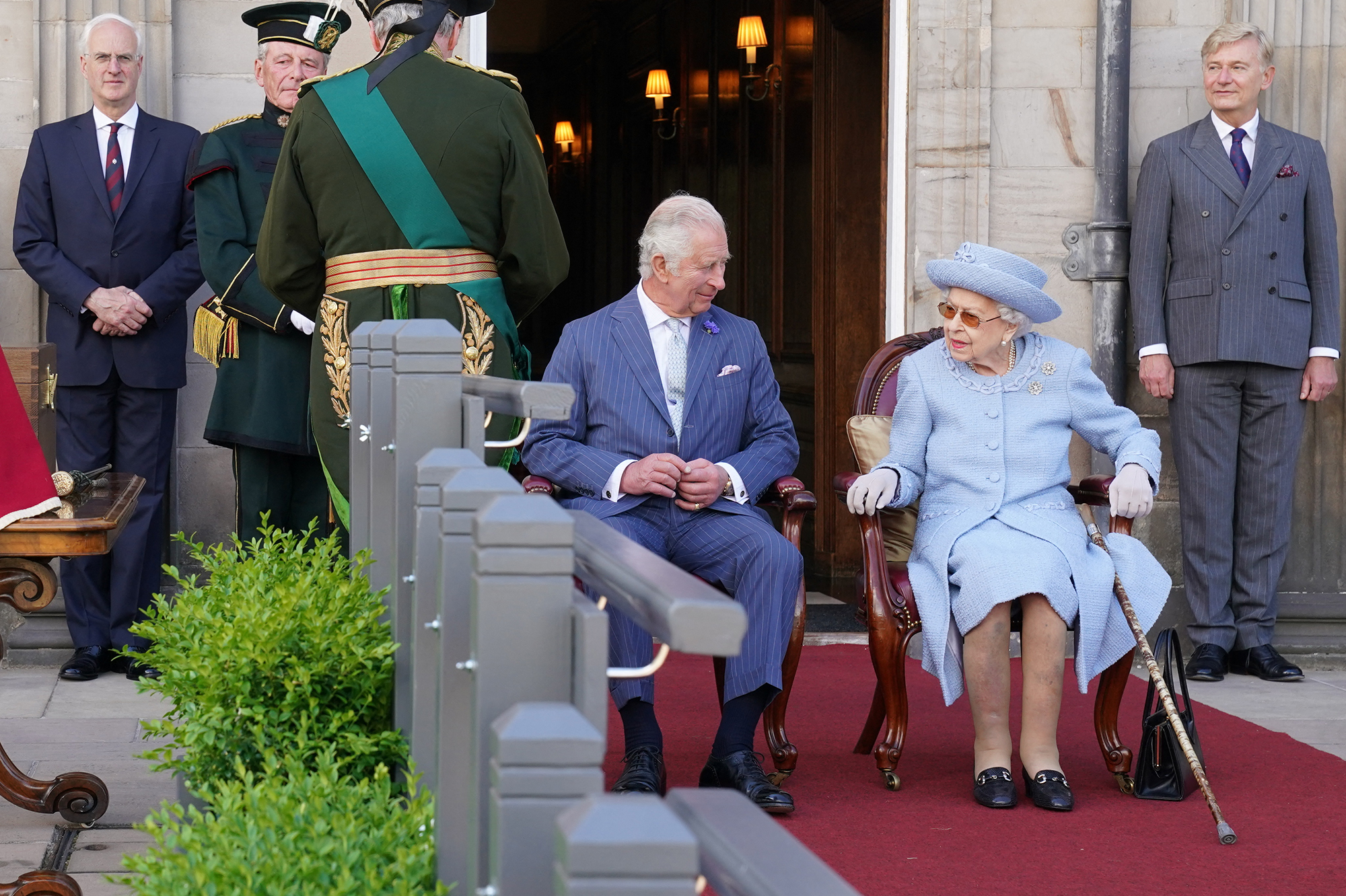 Charles and Queen Elizabeth seated in chairs on a red carpet outside a royal entrance in Scotland.