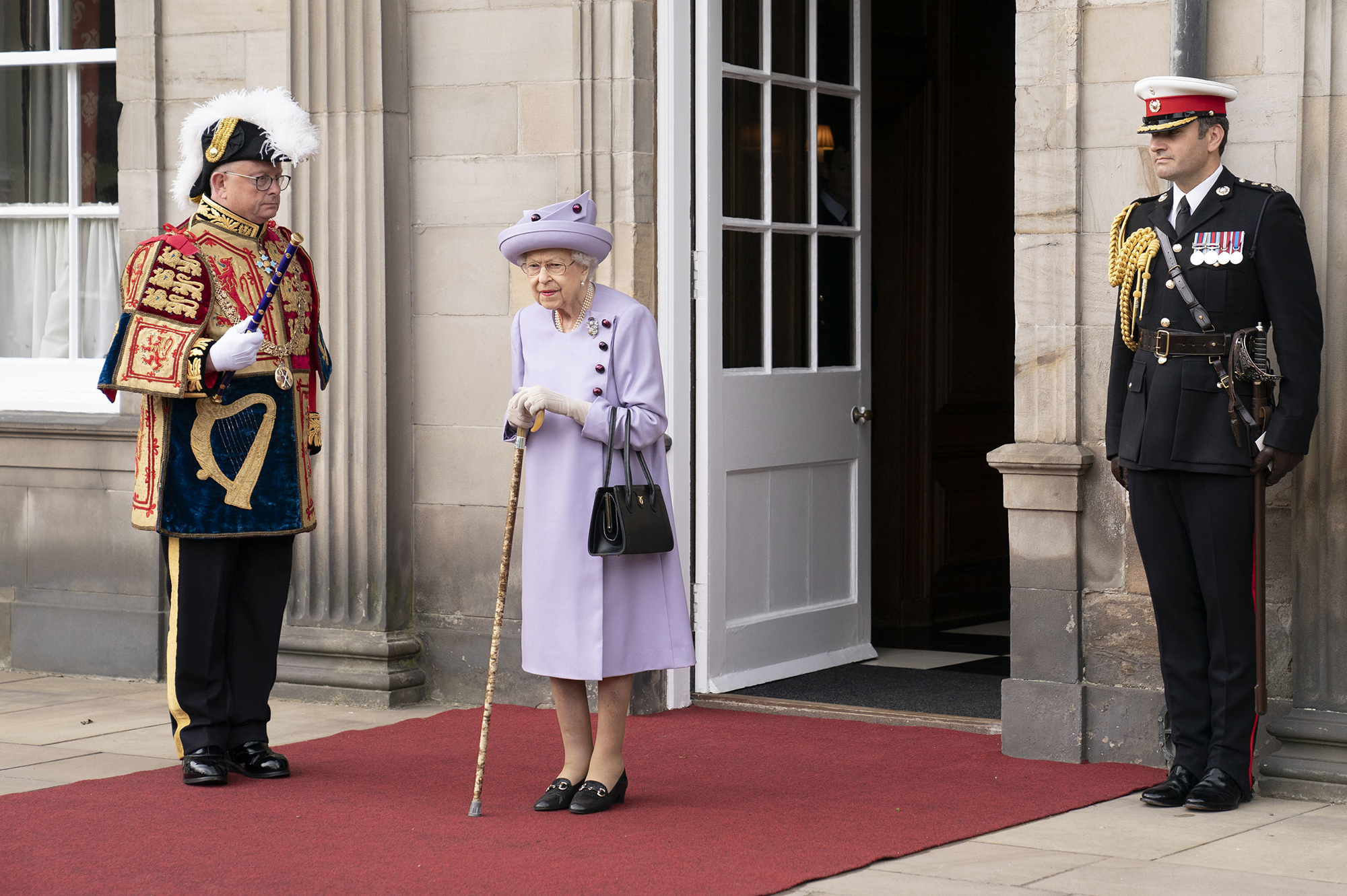 Queen Elizabeth standing on a red carpet holding a cane surrounded by two guards.