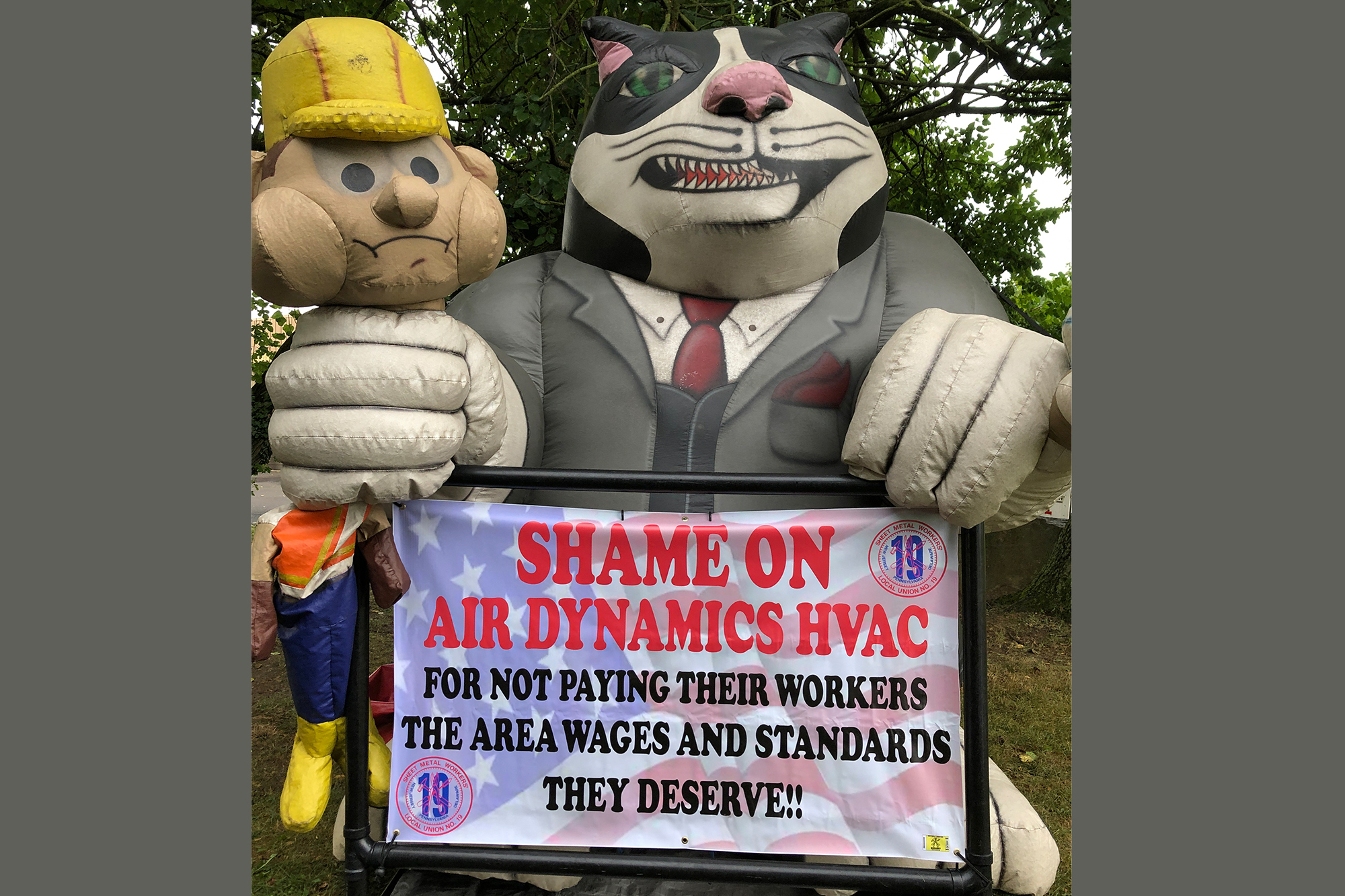 A large blow-up "fat cat" in a suit holds a construction-hatted worker in a stranglehold. The sign reads, "Shame on Air Dynamics HVAC for not paying their workers the area wages and standards they deserve!!"
