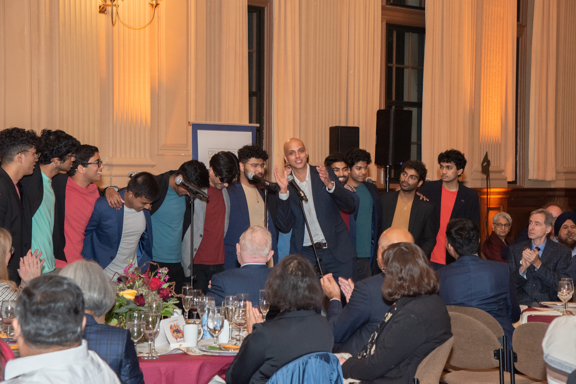 A group of men in colored shirts and dark blazers stands in front front of the microphones. An audience seated at circular banquet tables claps while Tariq Thachil thanks the group. 