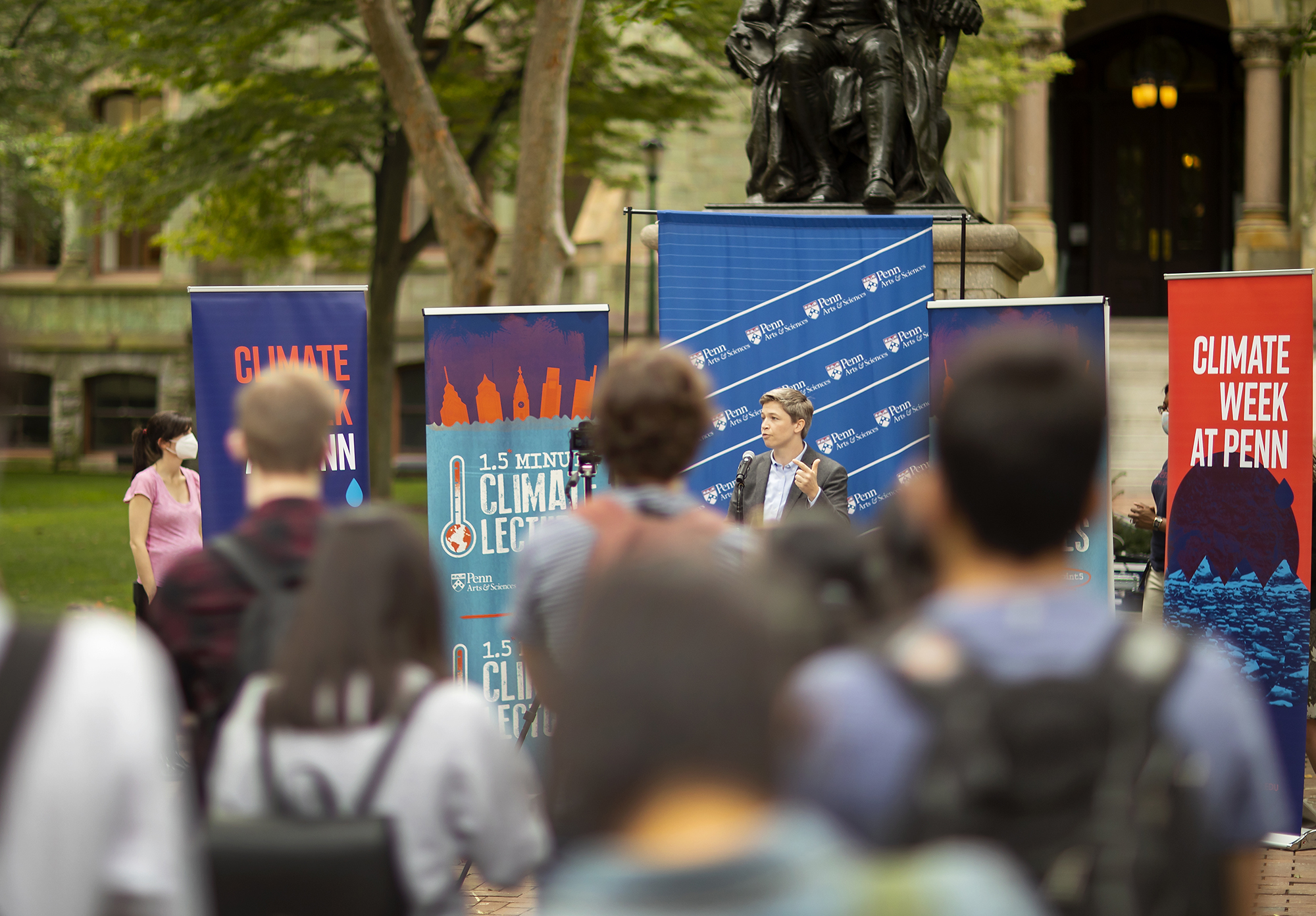 A person speaking at a lectern outside to a crowd with banners reading Climate Week at Penn