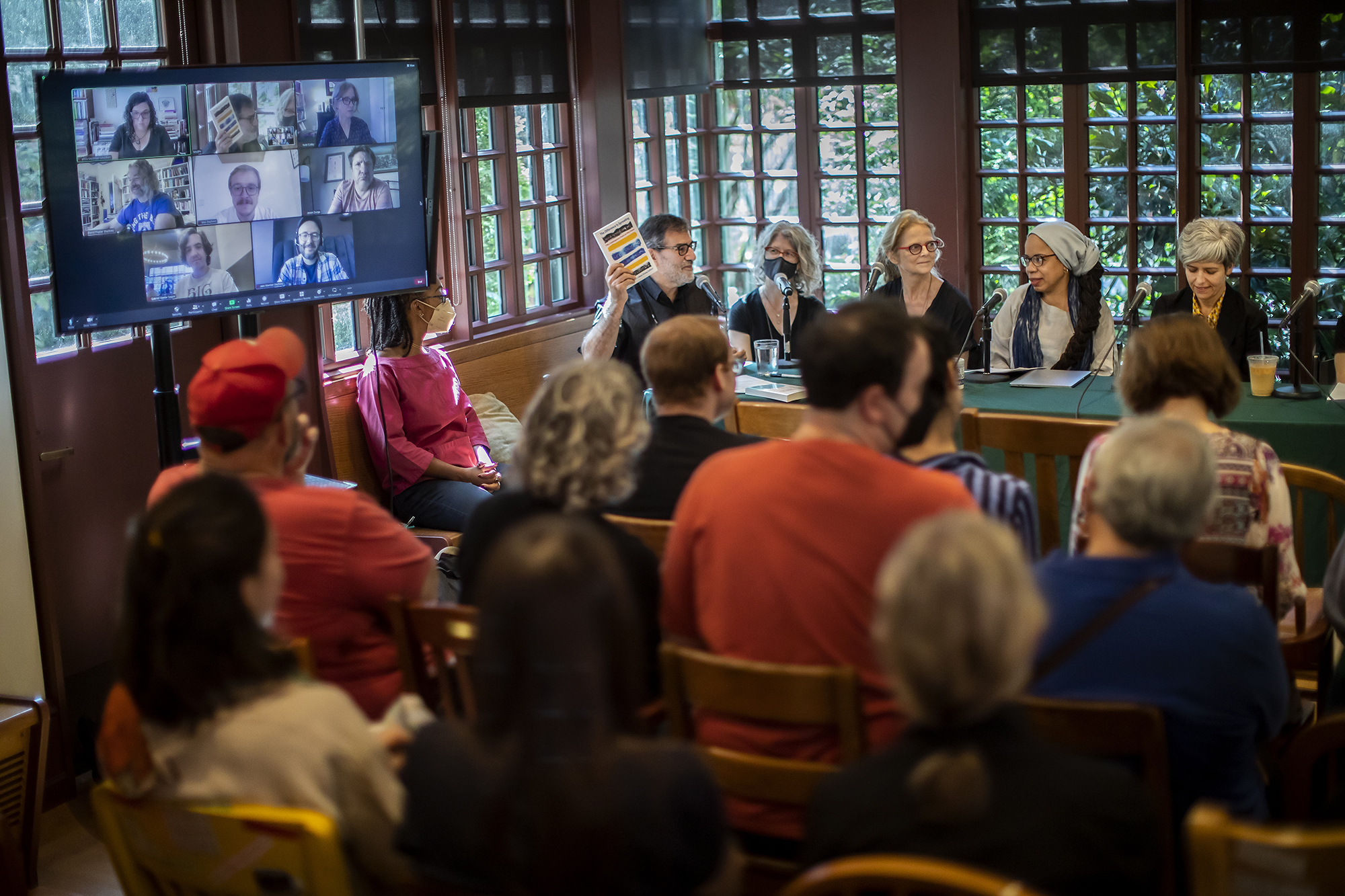 Al Filreis holds up the Book "The Difference is Spreading" while seated at a long table with four other people to his left and a television screen with the faces of several other people to his right in front of an audience