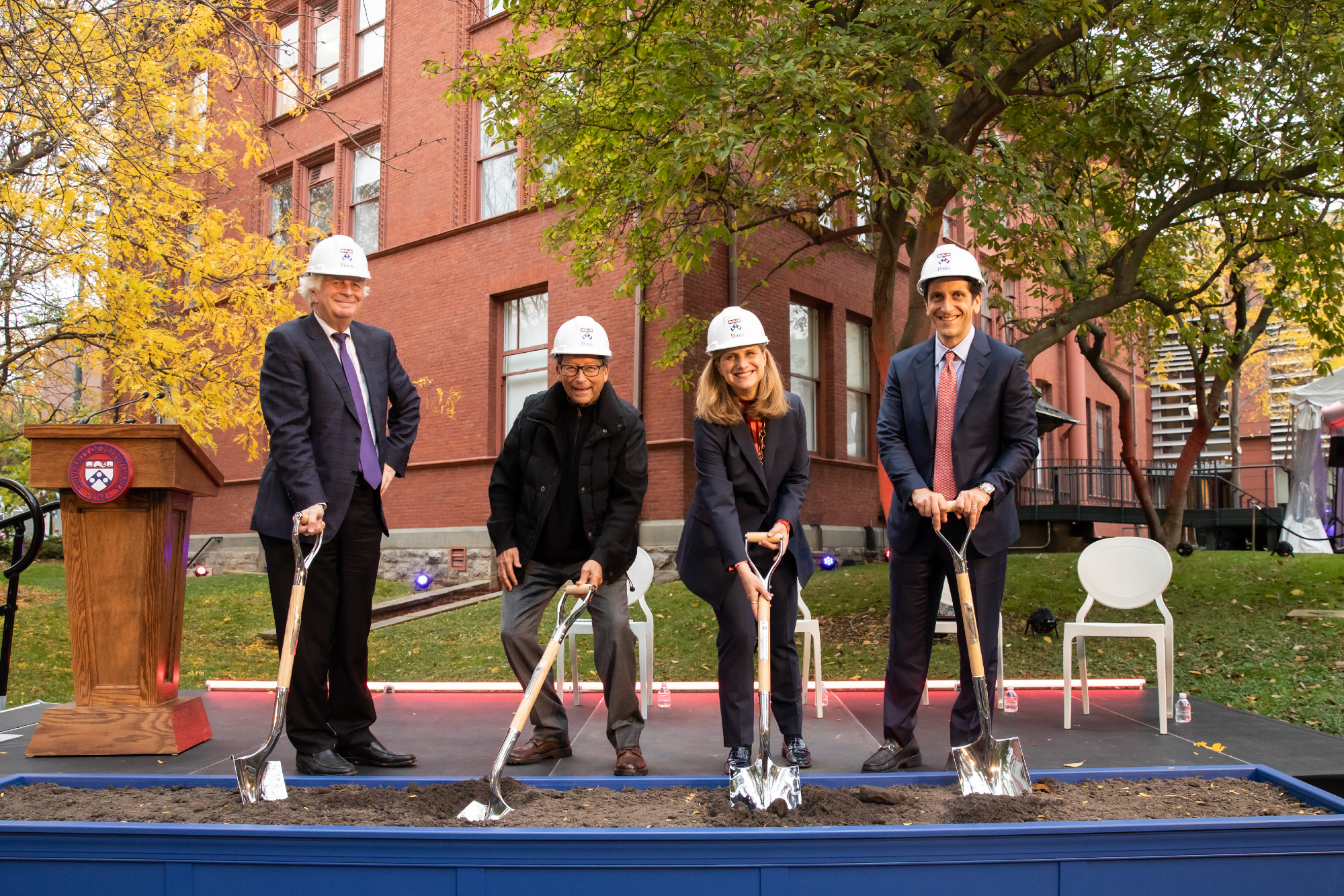 Four people wearing white hardhats are putting shovels into a trough of dirt in front of a stage, which is outside in front of a brick building. 