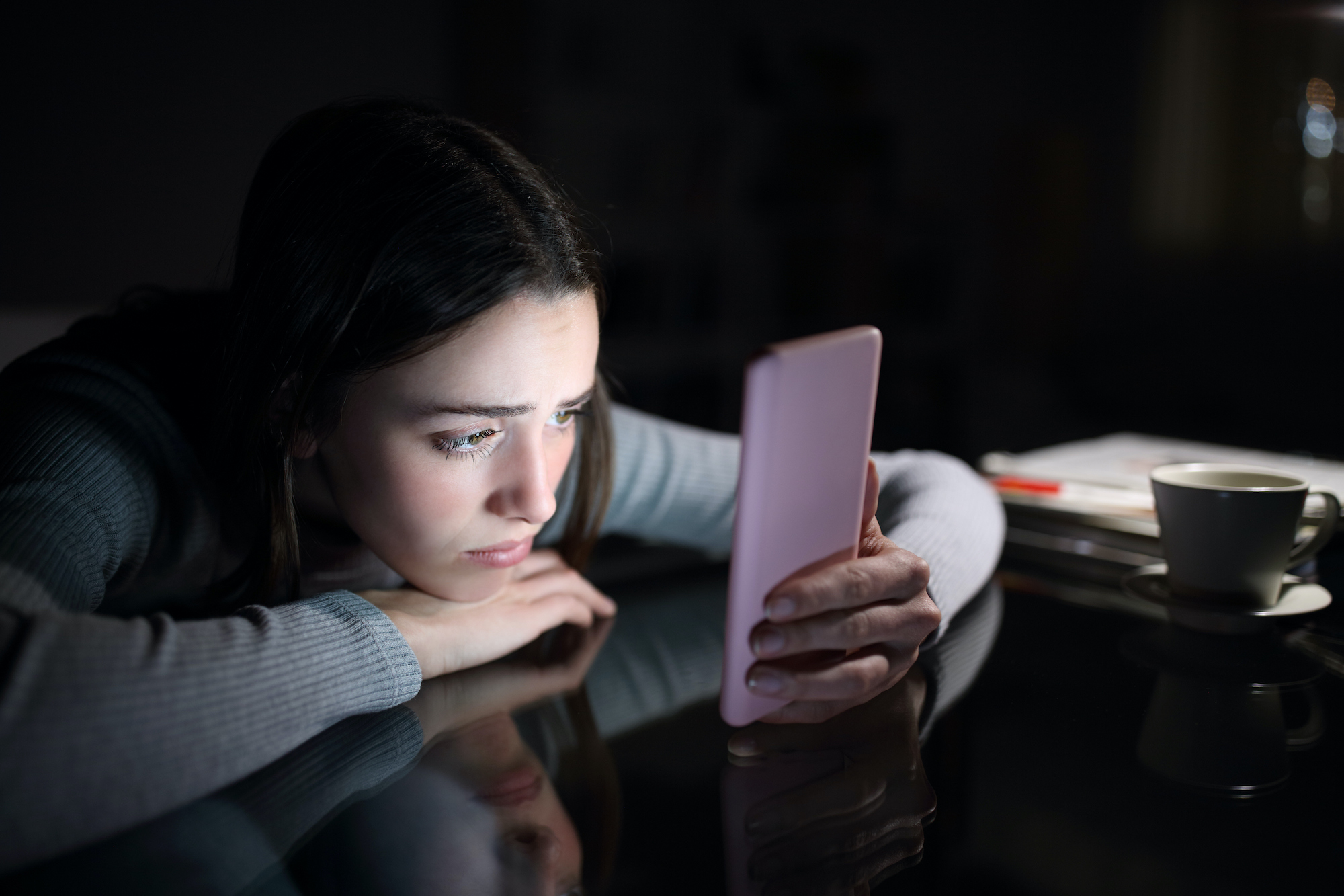 Person sitting in the dark, leaning on a desk, staring at a cell phone. A coffee cup and pile of papers sit nearby.