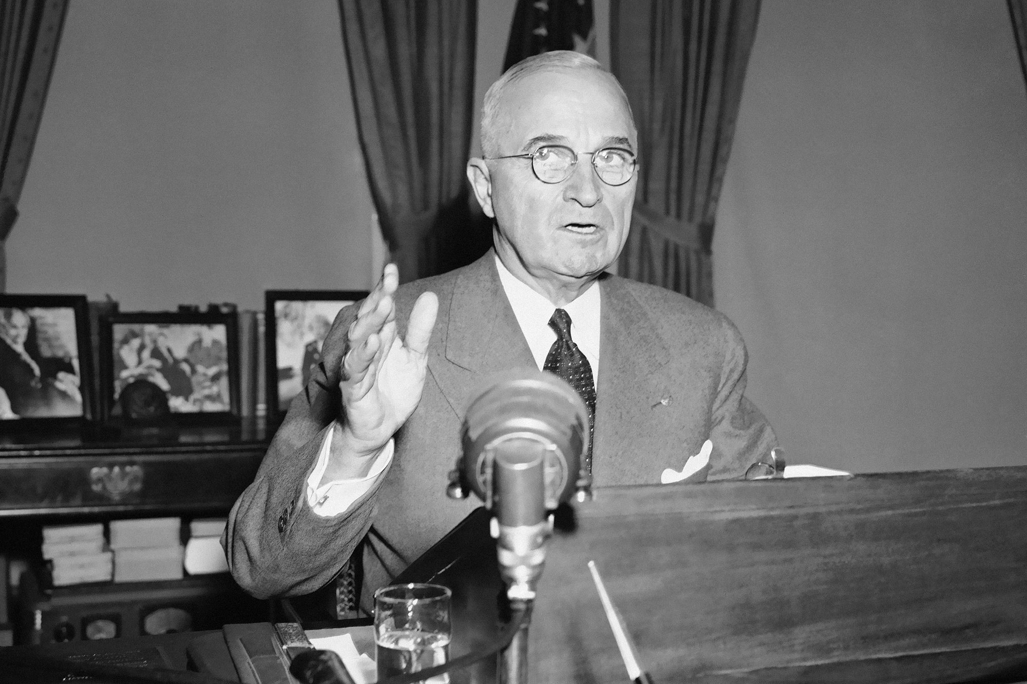 A black and white image of Harry Truman speaking in front of a microphone. 