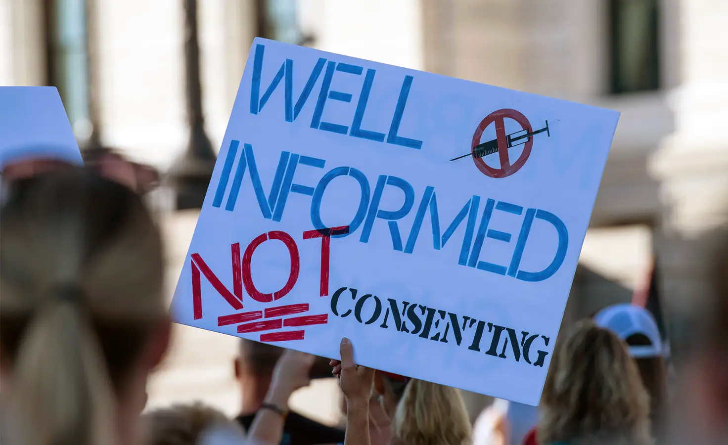 A person holds a sign during a protest that reads WELL INFORMED NOT CONSENTING.