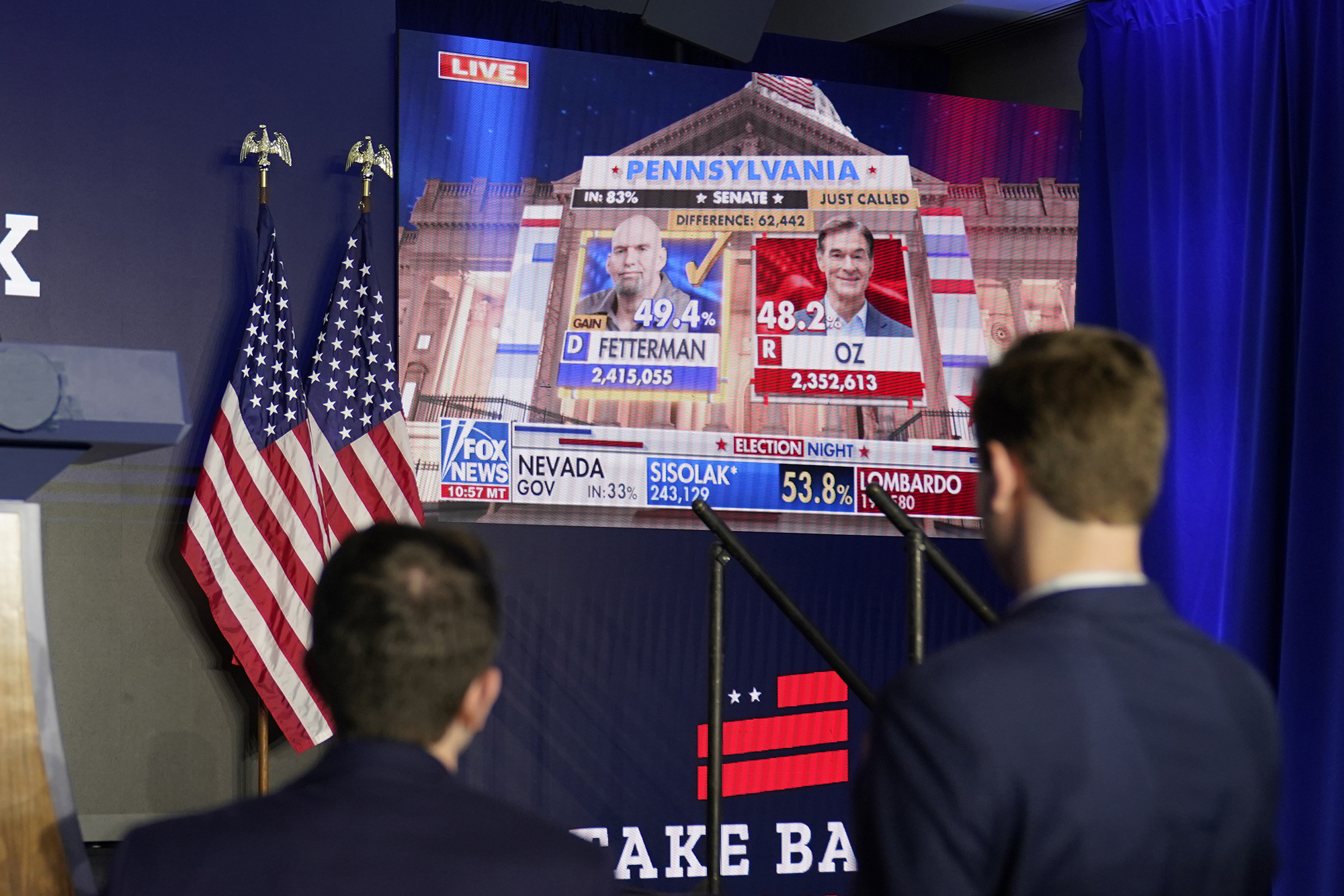 Two men in suits face a large TV screen showing Pennsylvania 2022 Senate election results.