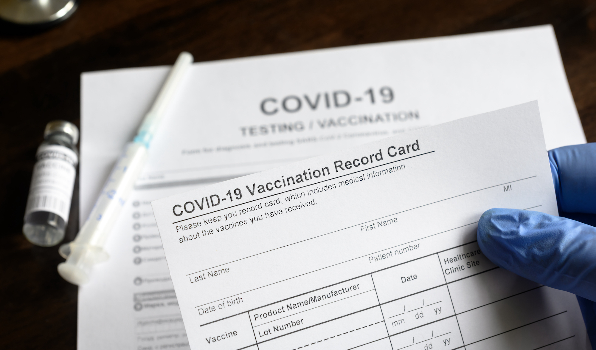 A gloved hand holds a COVID-19 Vaccination Record card. Blurred in the background are a closed needle, a vial of medication, and a sheet of paper that reads "COVID-19 Testing/Vaccination." 
