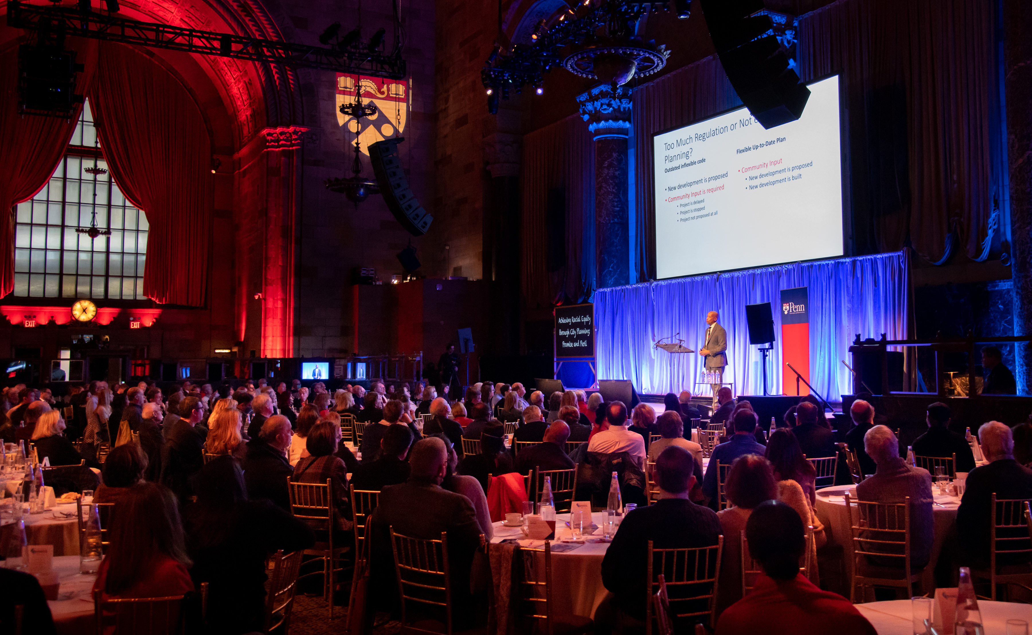 Lance Freeman, at a podium on a stage, speaks to a crowd with a presentation displayed in the background