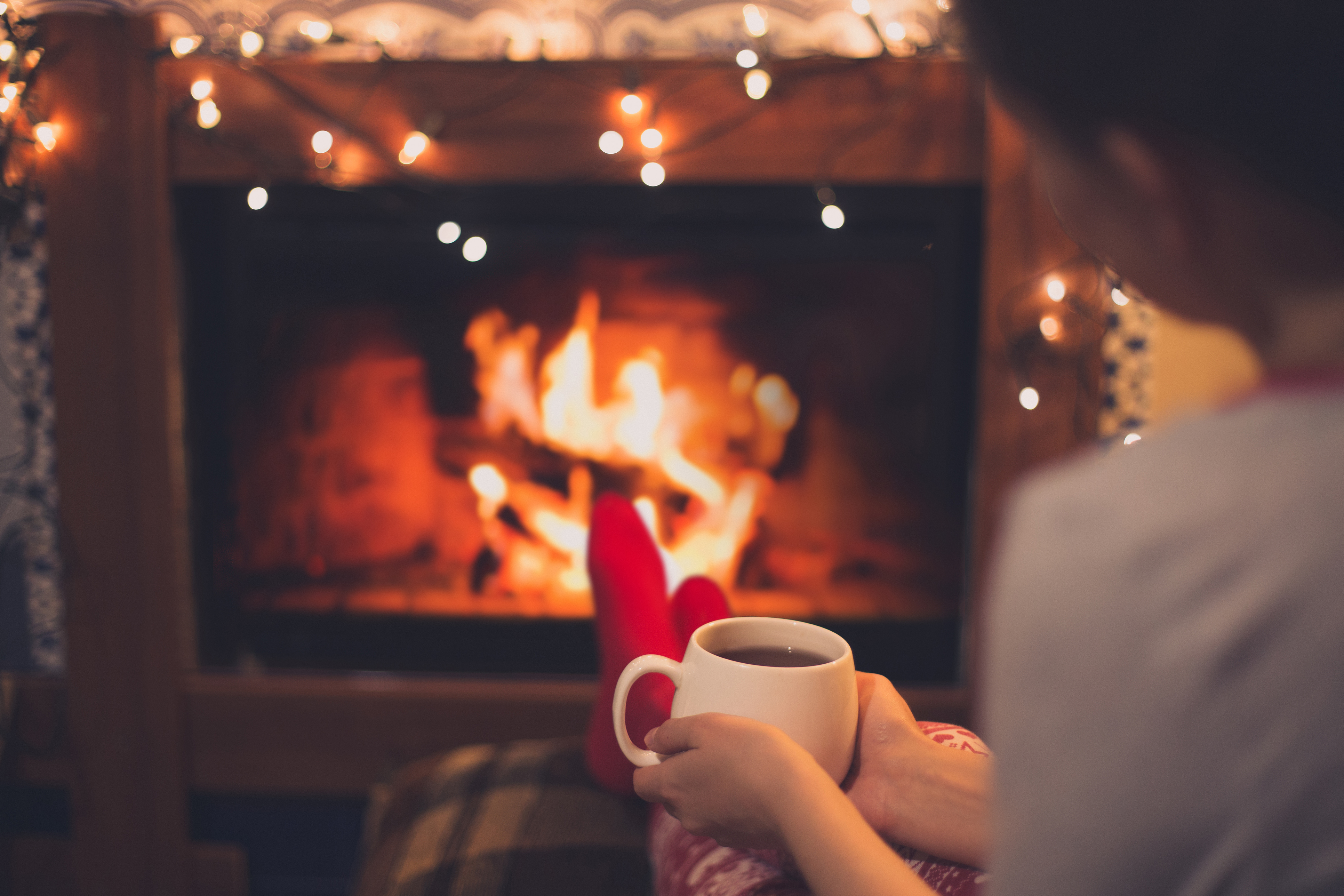 A woman sits by a fireplace with twinkle lights and drinks tea