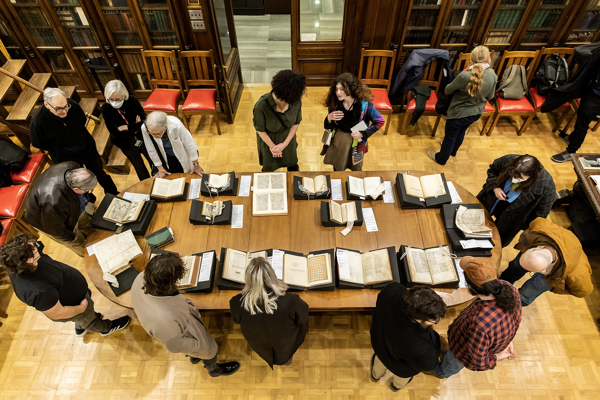 An overhead view of a long table in a historic library room with several open texts 
