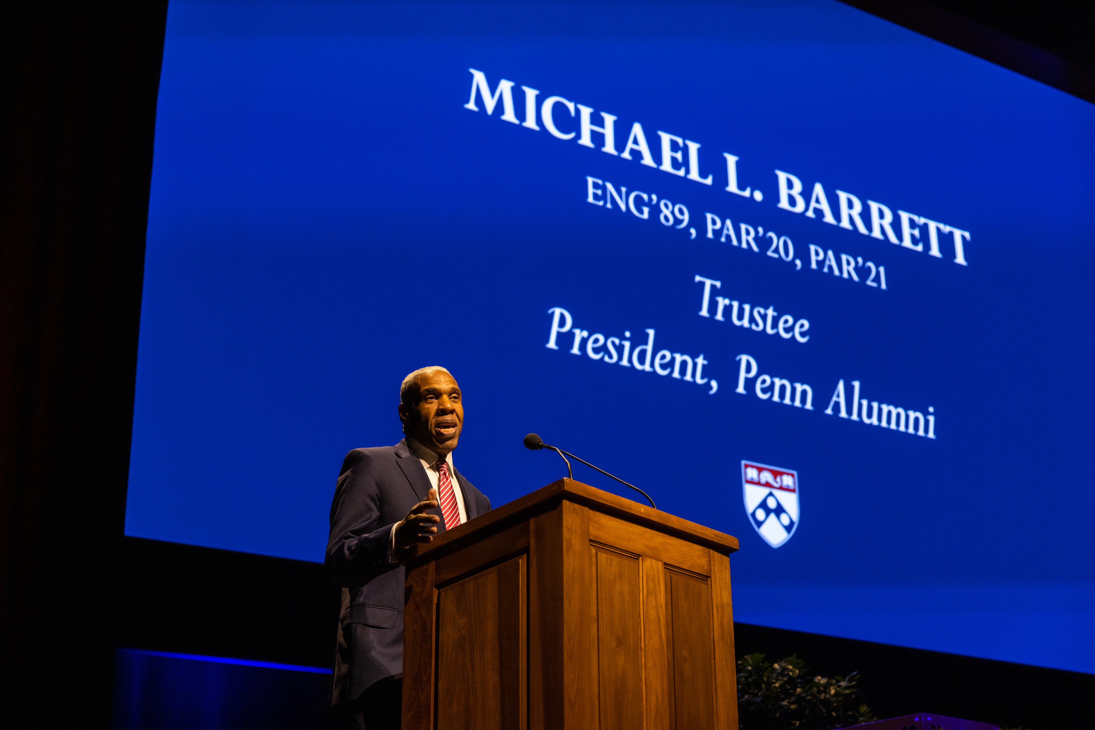 Michael Barrett speaking at a podium with the words Michael L. Barrett Eng’89, Par’20, Par’21, Trustee, President, Penn Alumni on a screen behind him. 
