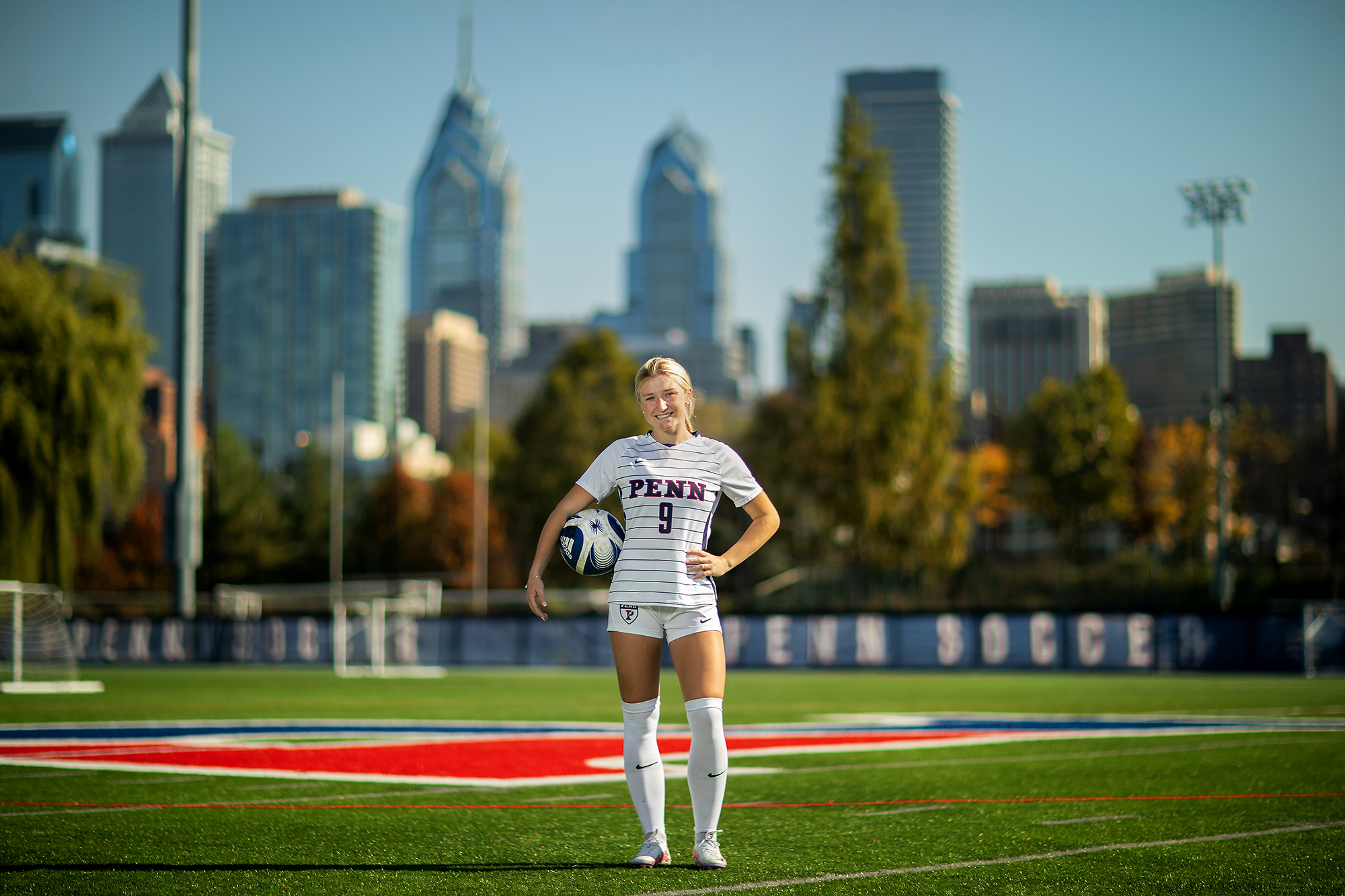 Lawton stands confidentally in the soccer field with her hand on one hip and the other draped over a soccer ball in front of the philadelphia skyline.