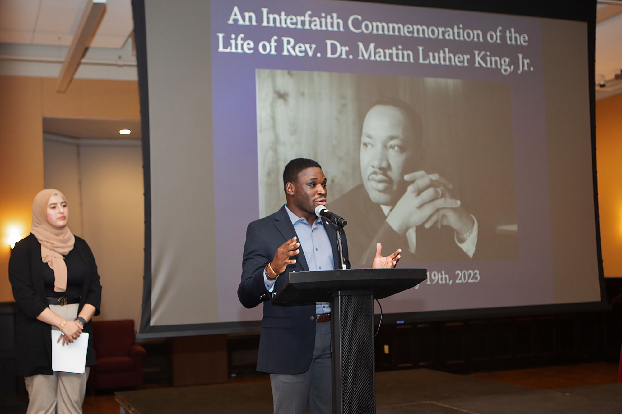 A man speaks at a podium with a woman waiting behind him. The screen says "An Interfaith Commemoration of the Life of Rev. Dr. Martin Luther King, Jr."