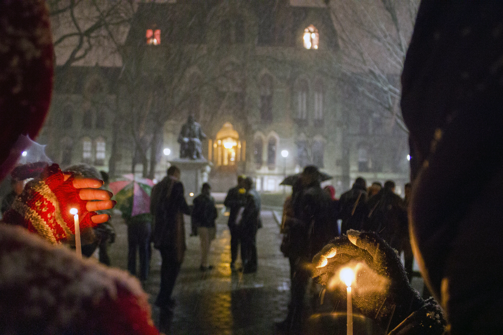 Walkers cup lit candles with mittened hands as snow falls on College Hall