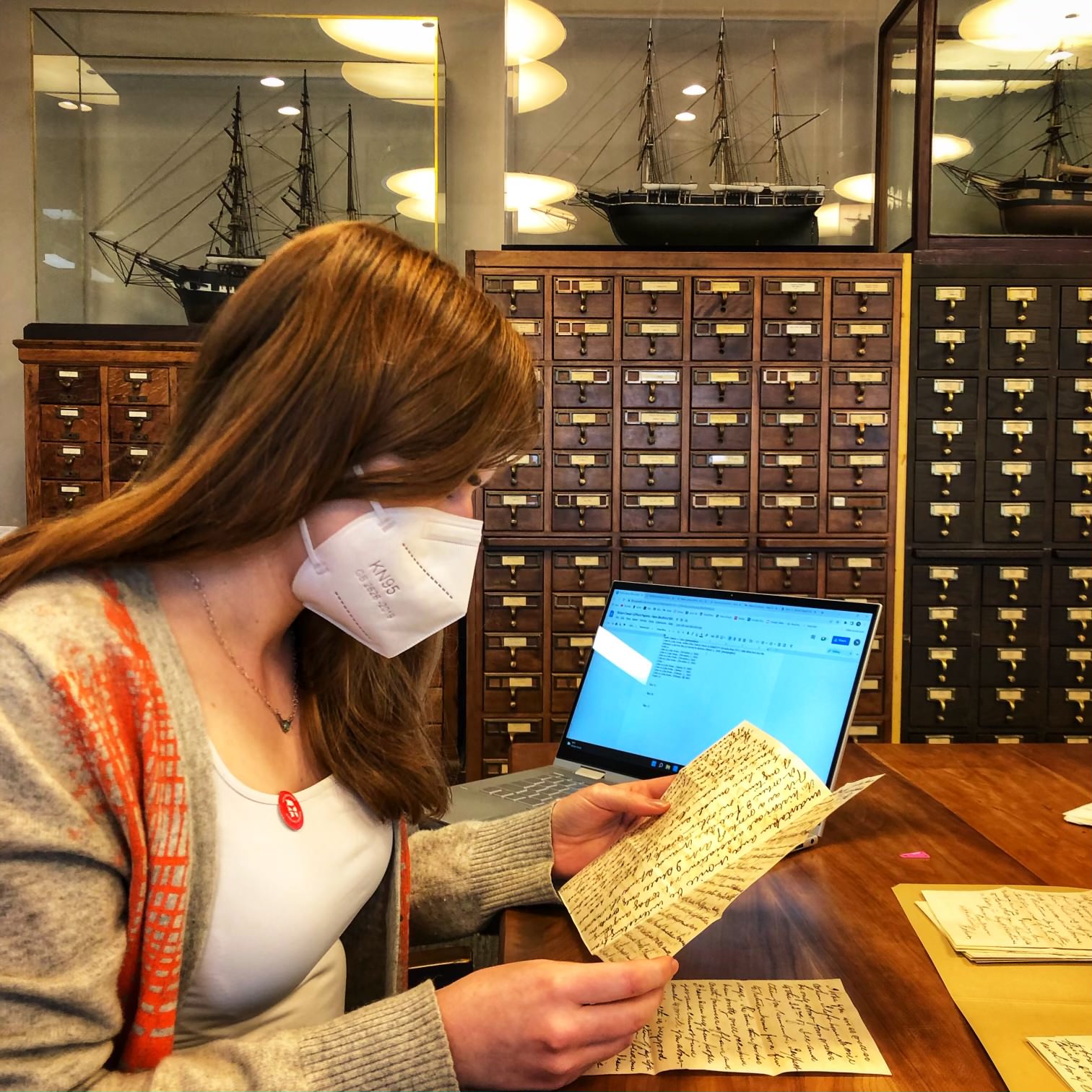 Aili Waller reading a document with hand-written text at a table in a library with card catalogues behind her. 
