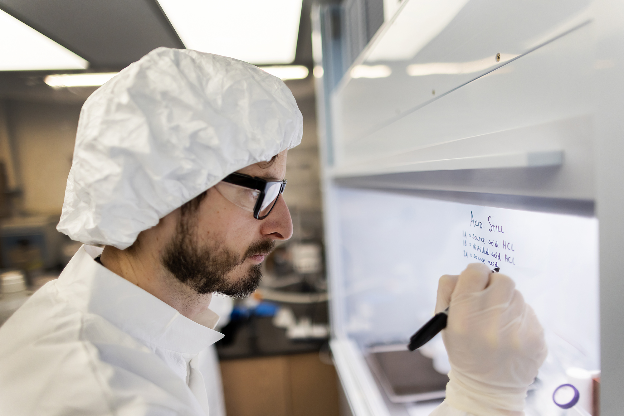 Scientist with protective cap, glasses, gloves, and jacket writes on a glass laboratory hood