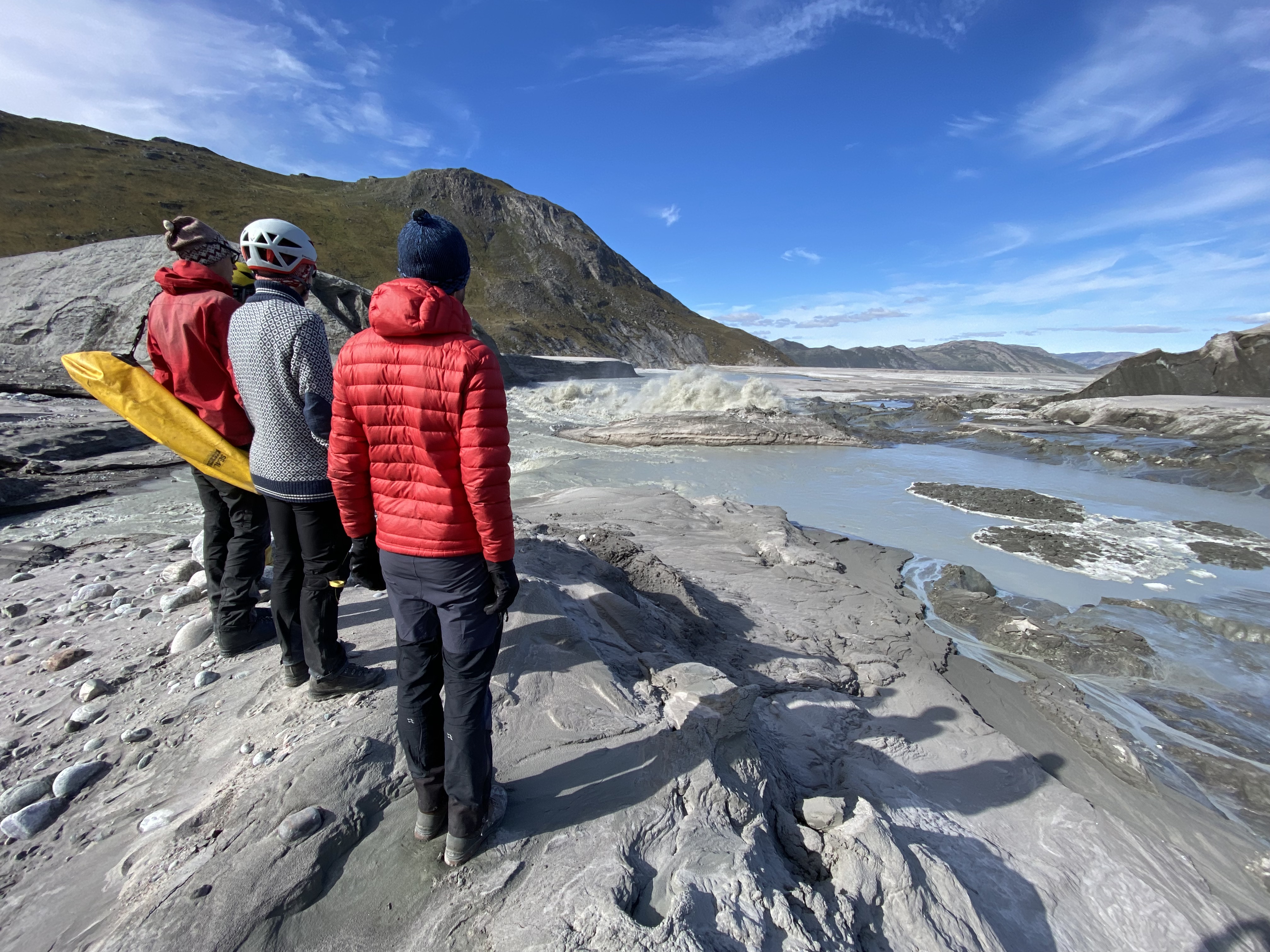Three scientists observe glacier meltwater beneath mountains and clear skies