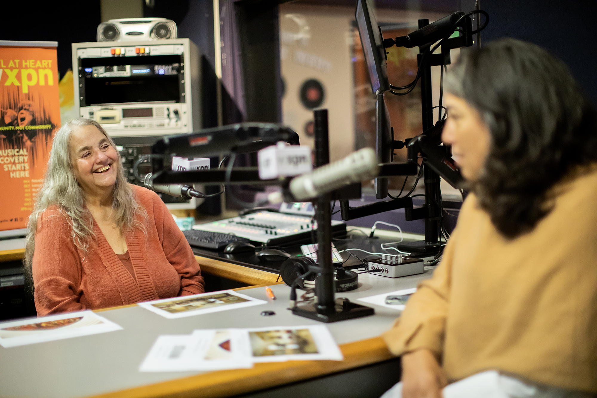 Kathy O'Connell laughs while speaking with Gwendolyn DuBois Shaw while they sit in front of microphones in the radio recording studion. 