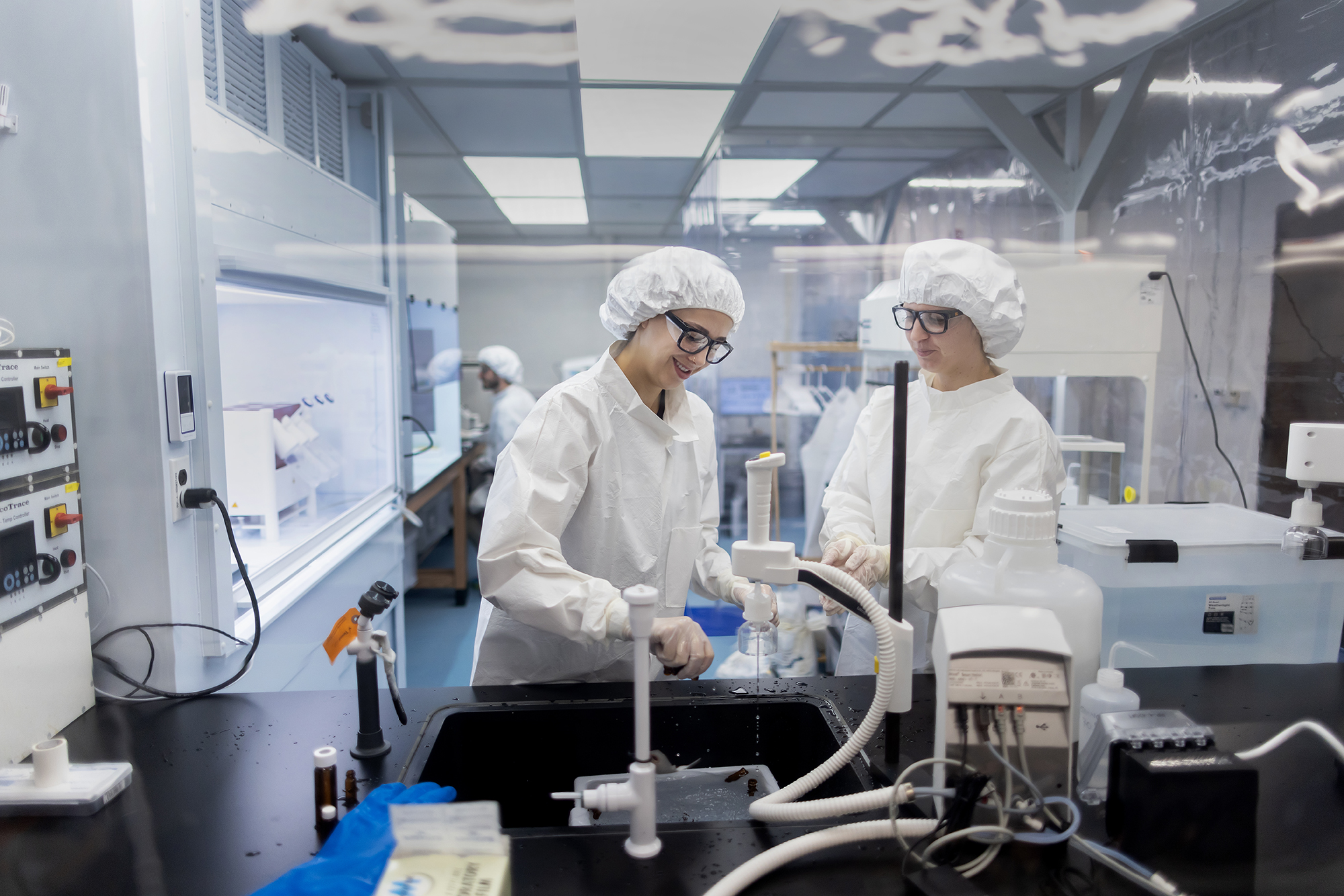 Two scientists in protective gown, cap, gloves, and glasses work in a lab cleaning equipment