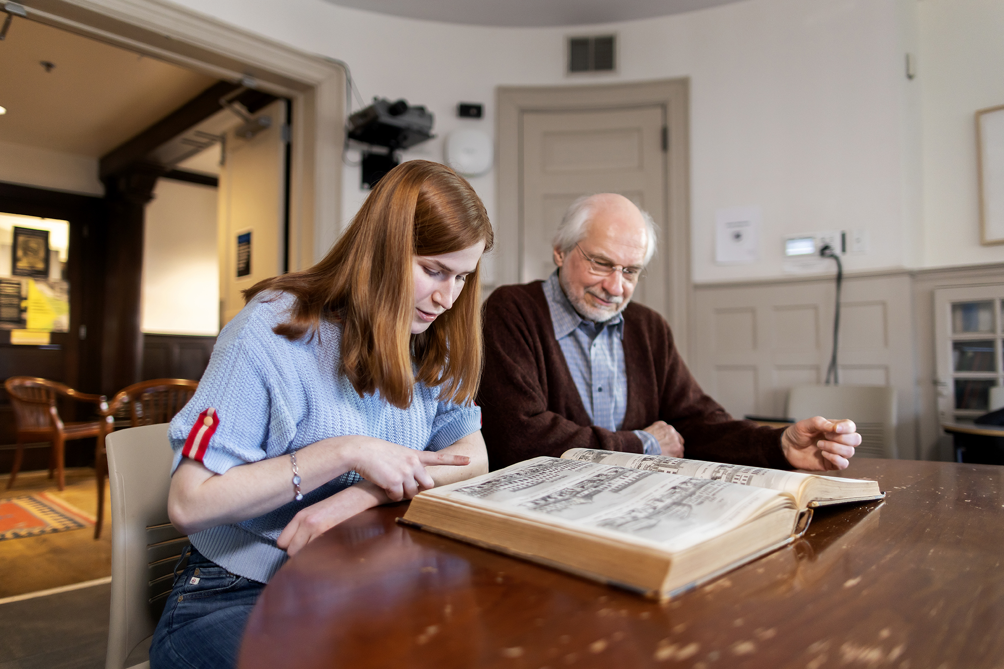 Aili Waller and Professor Michael Leja looking at a historic book while sitting at a wooden table in a historic room. 