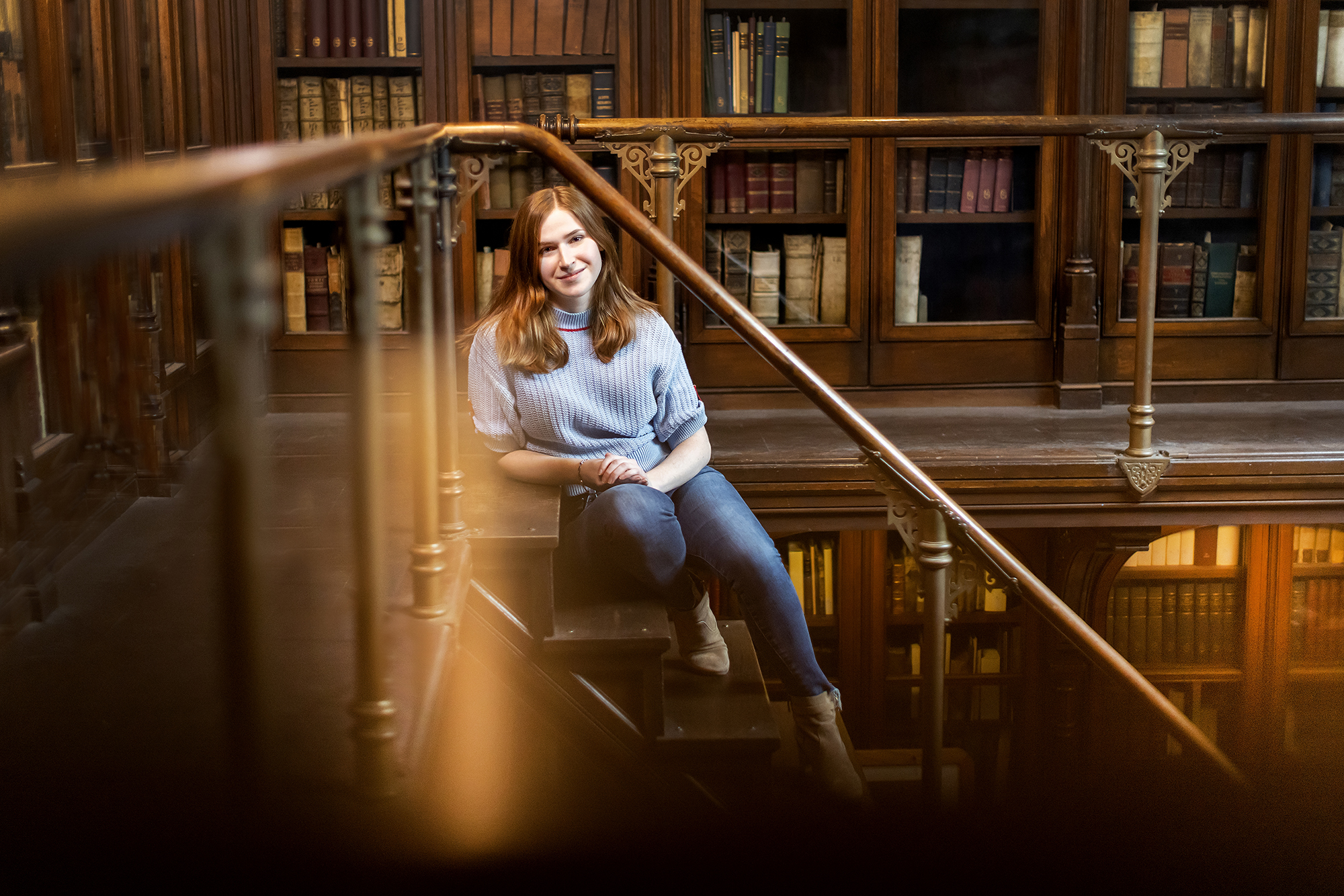 Aili Waller sitting on the stairway in a historic library with books behind her in glass-doored shelving. 