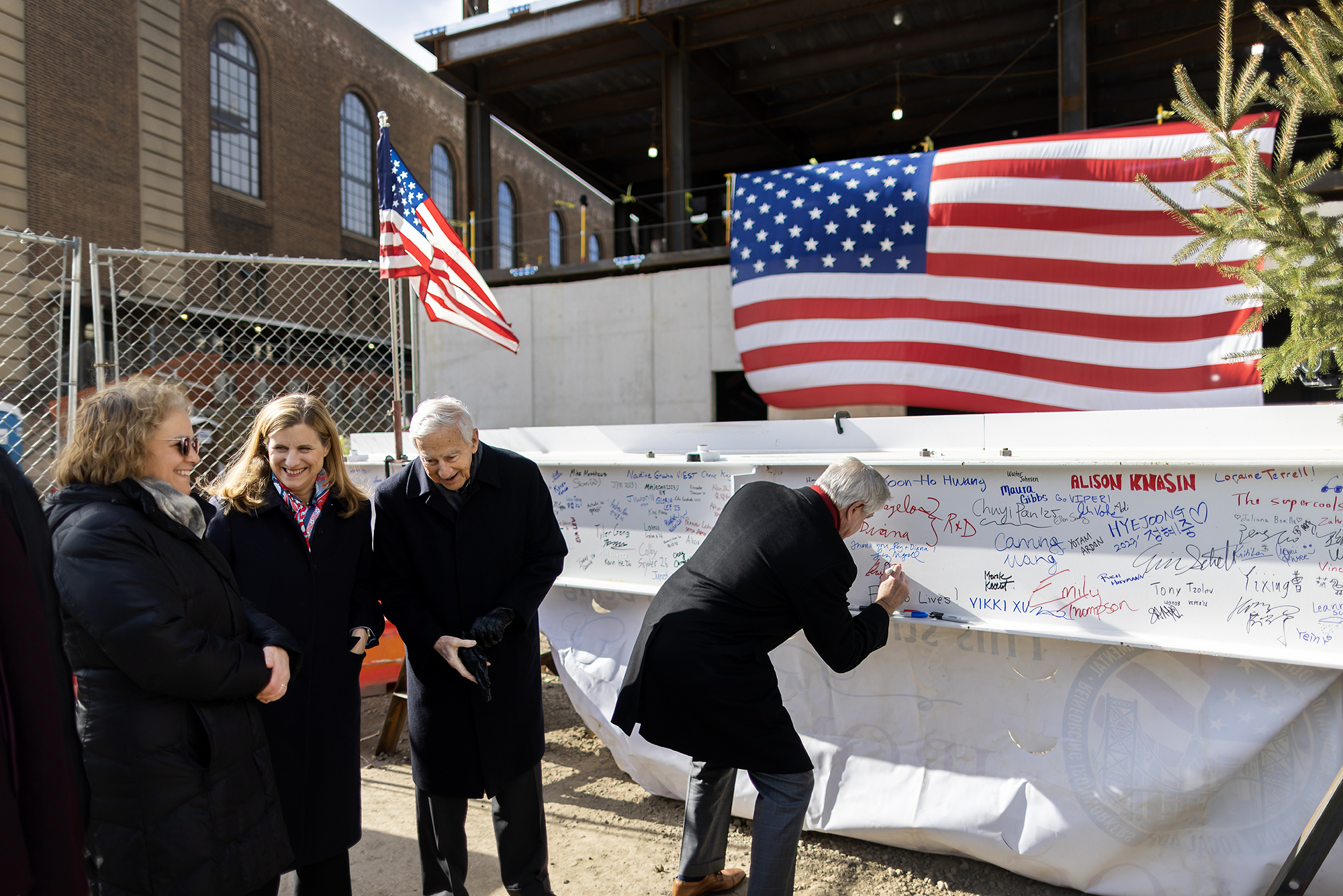 Penn President Liz Magill, Mr. Vagelos, and another person stand on the sidewalk while a fourth person signs the final beam.