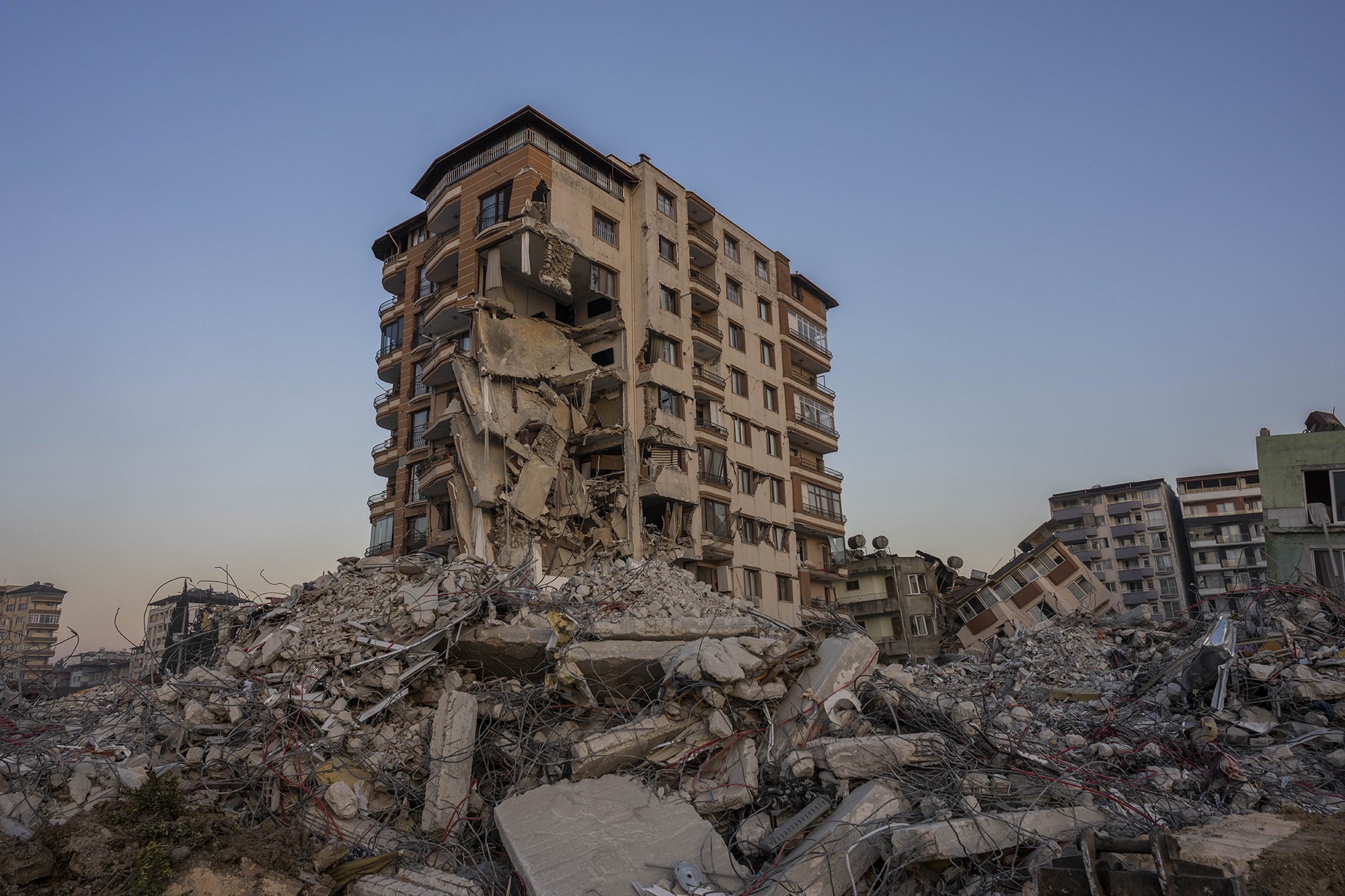 A building standing amid piles of rubble set against a blue sky. The building is partially collapsed.