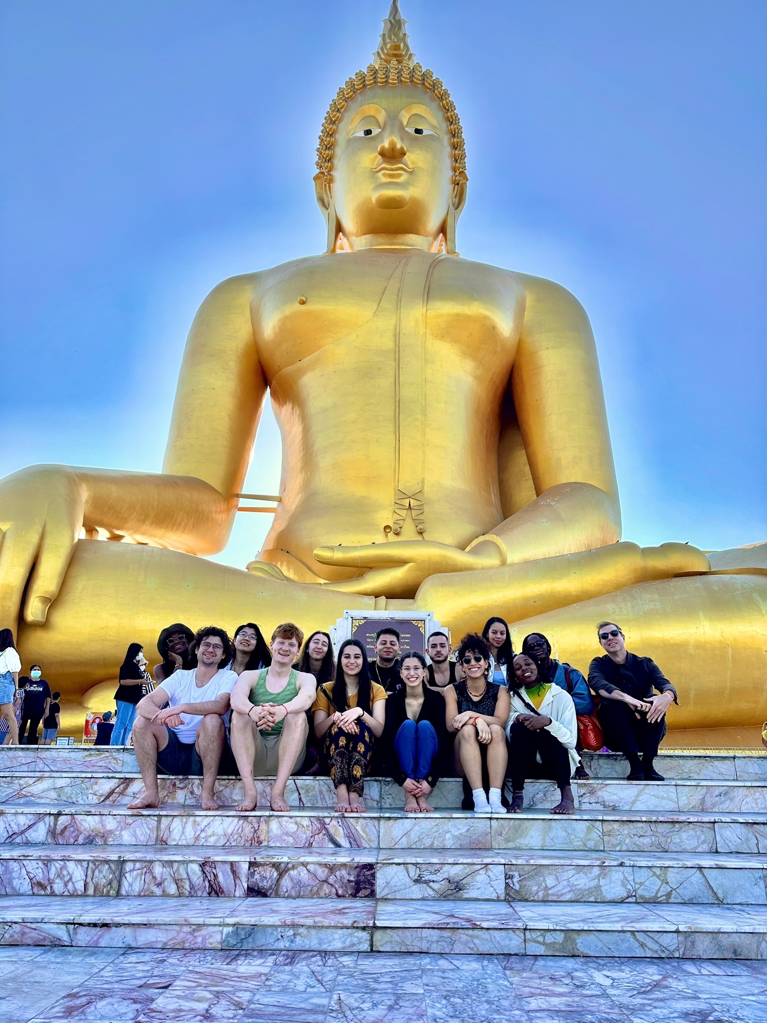 A group of students sit on the stone steps beneath a giant golden Buddha