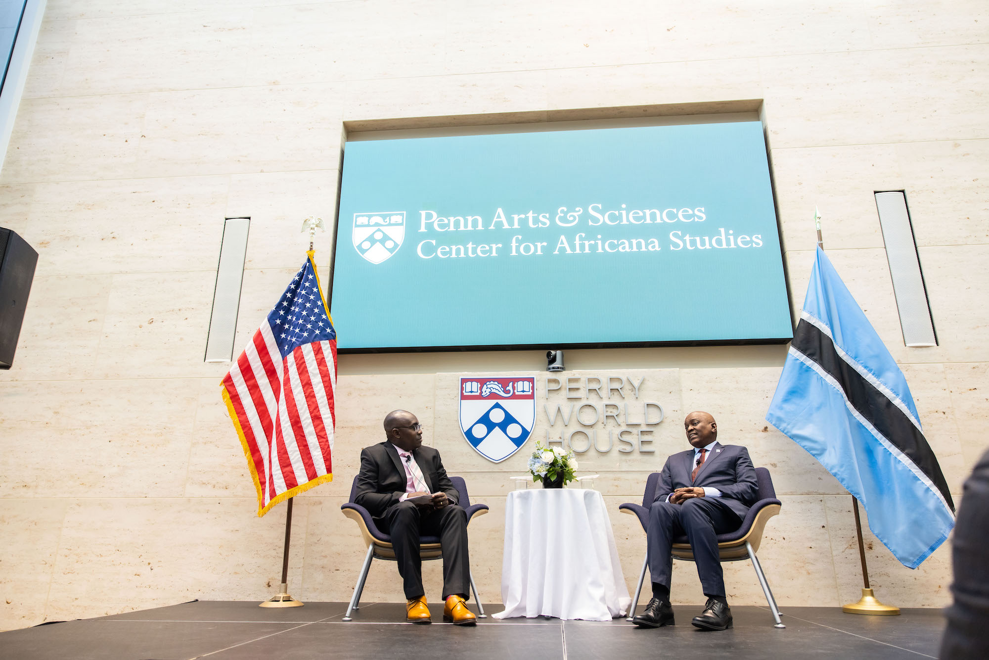 Two people sit in chairs on a stage between an American flag and a flag of the Republic of Botswana, in front of a sign reading Penn Arts and Sciences Center for Africana Studies
