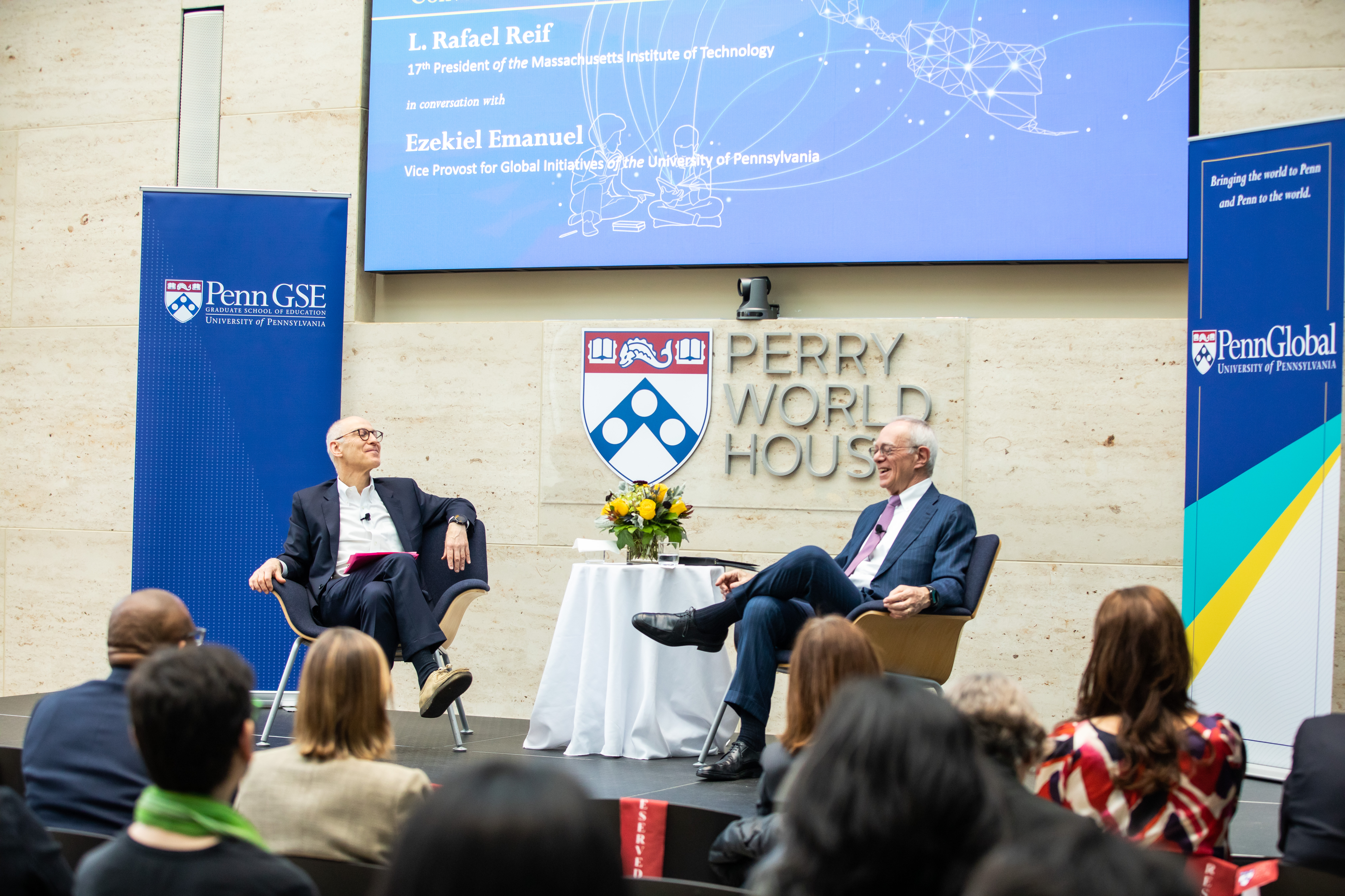 two people sitting on a stage with Perry World House on wall behind them