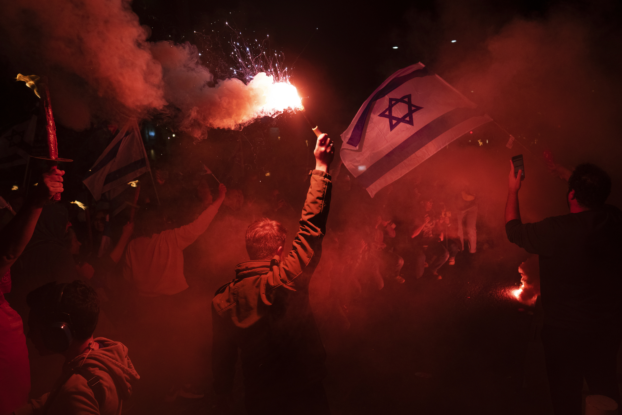 An Israeli protester holds a lit flare giving off a red glow as another waves an Israeli flag in a nighttime protest.